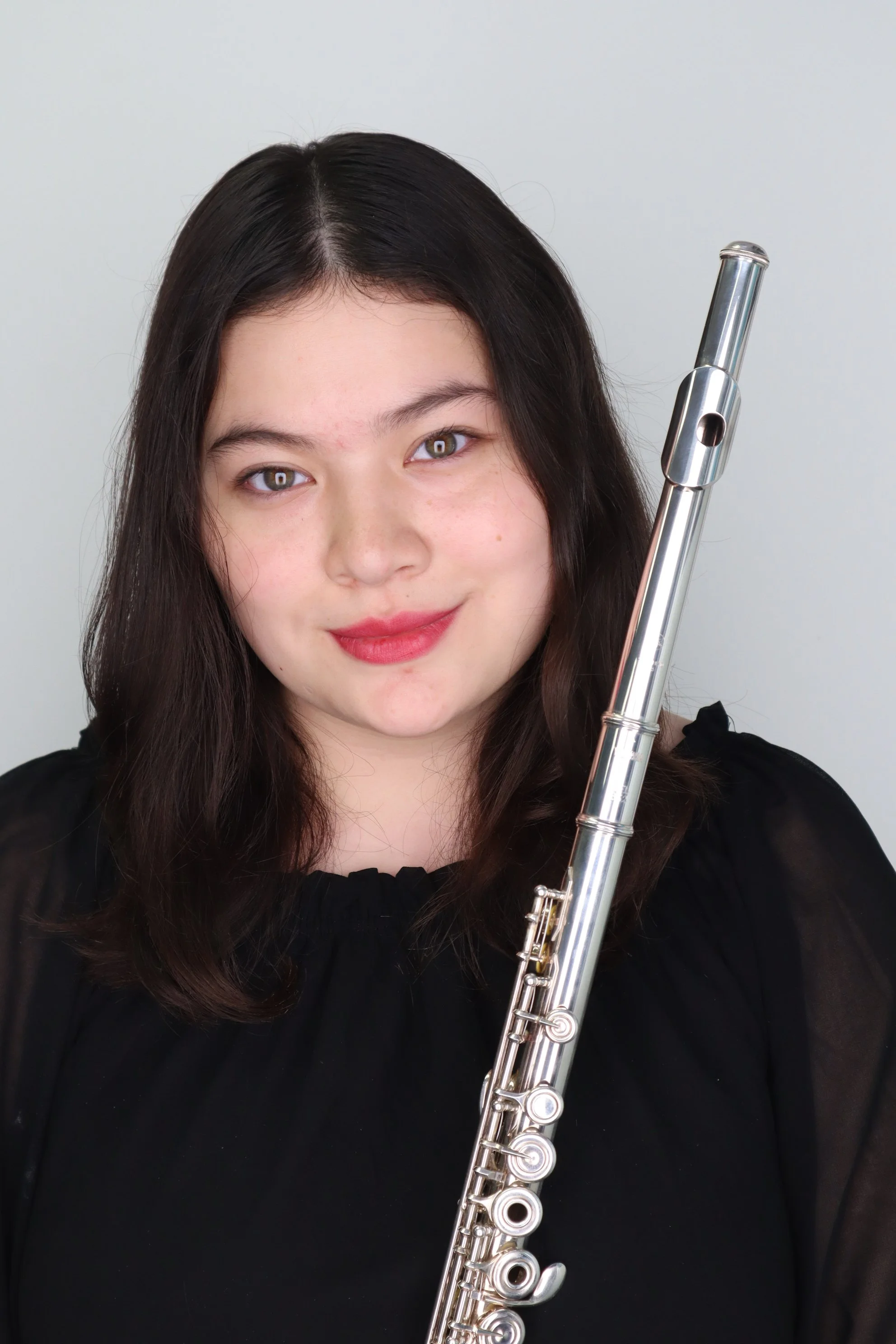 Young woman with dark brown hair holding a silver flute, wearing a black top, smiling, against a plain light background.