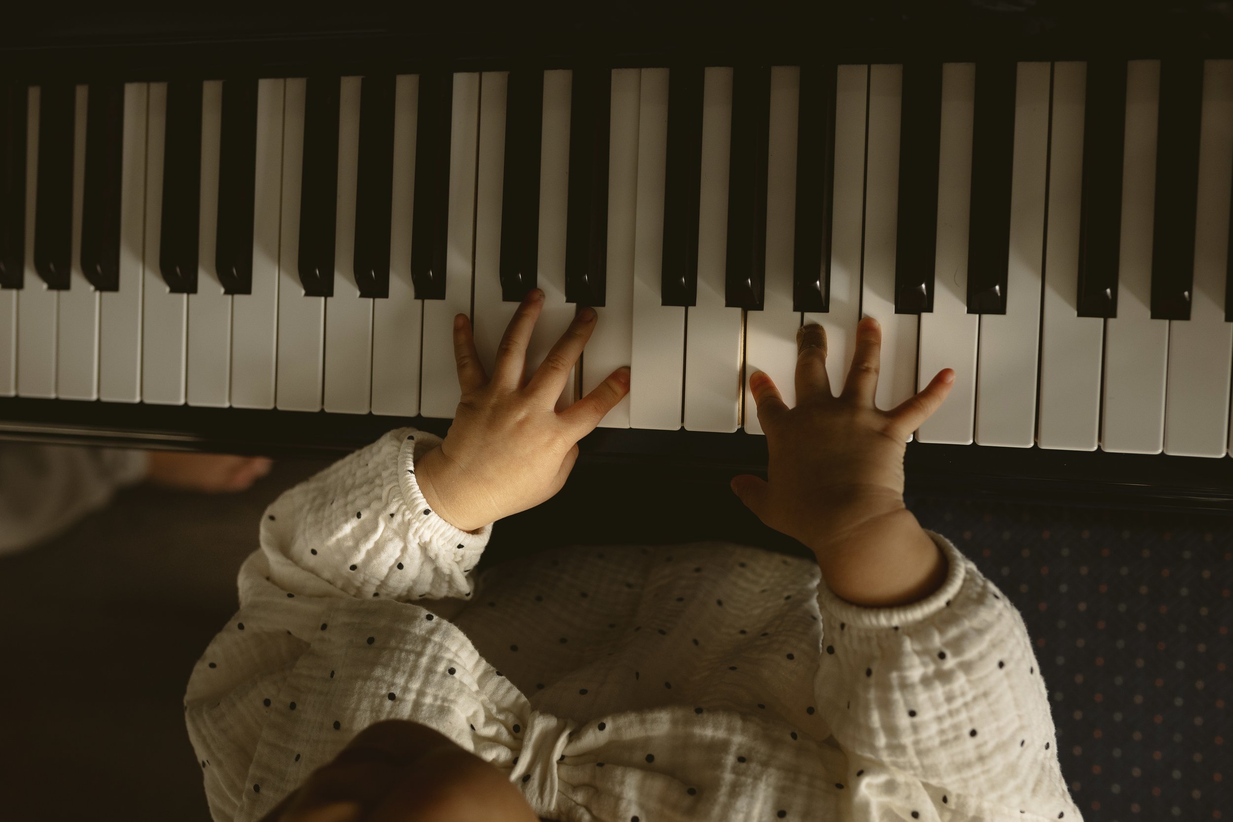 Close-up of a young child's hands playing a black and white piano keyboard, wearing a white polka dot long-sleeve shirt.