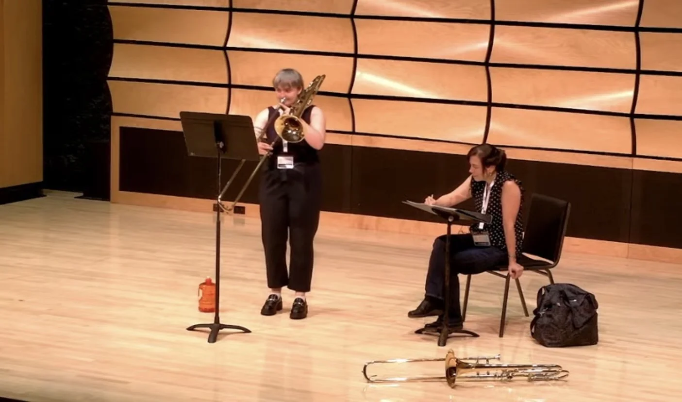Two women performing music on stage: one playing a saxophone, and the other seated with a music stand and a trumpet on the floor. The stage has a wooden background with black accents.