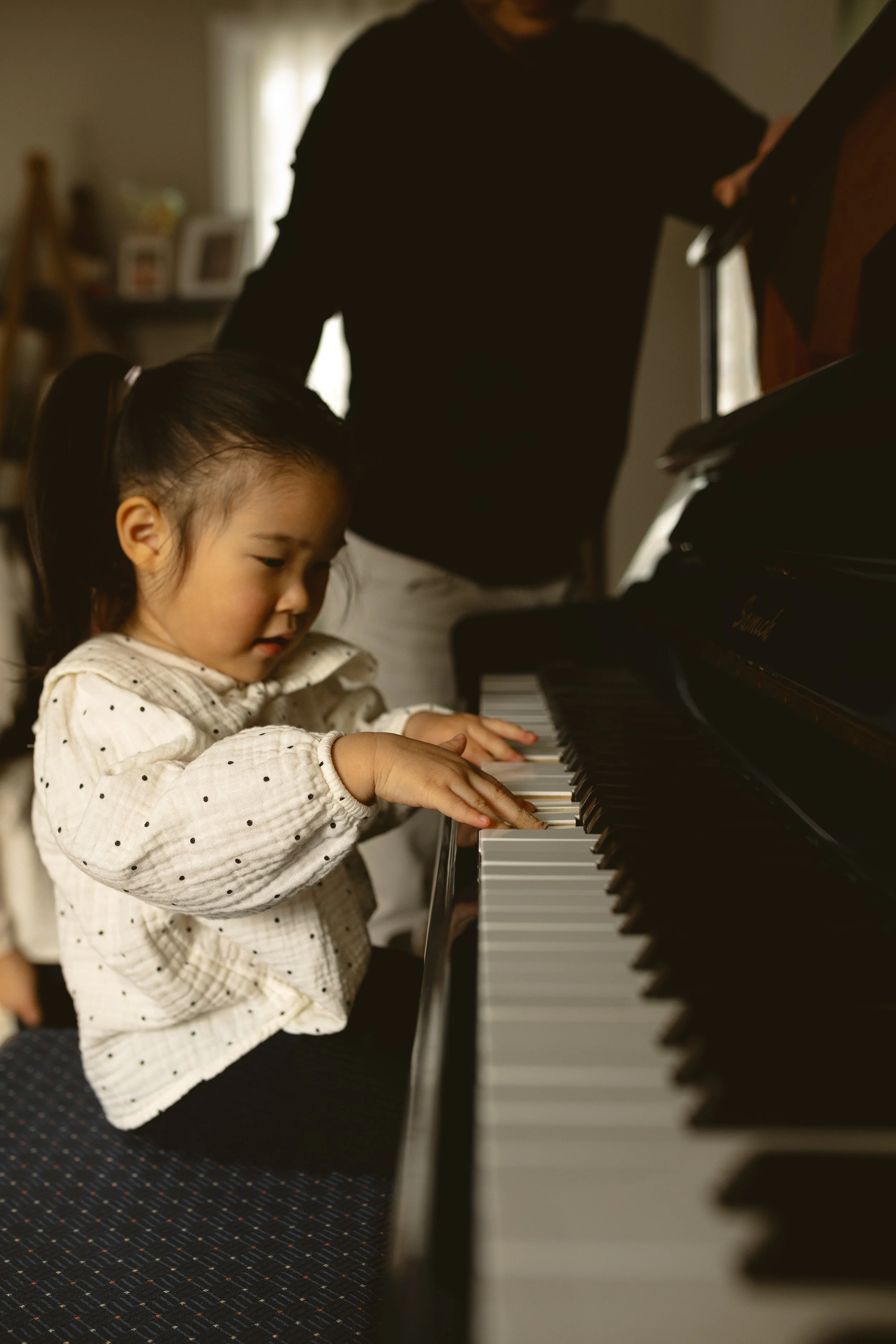 A young girl playing the piano with a person standing beside her, possibly giving guidance, in a cozy room with soft lighting and a patterned carpet.