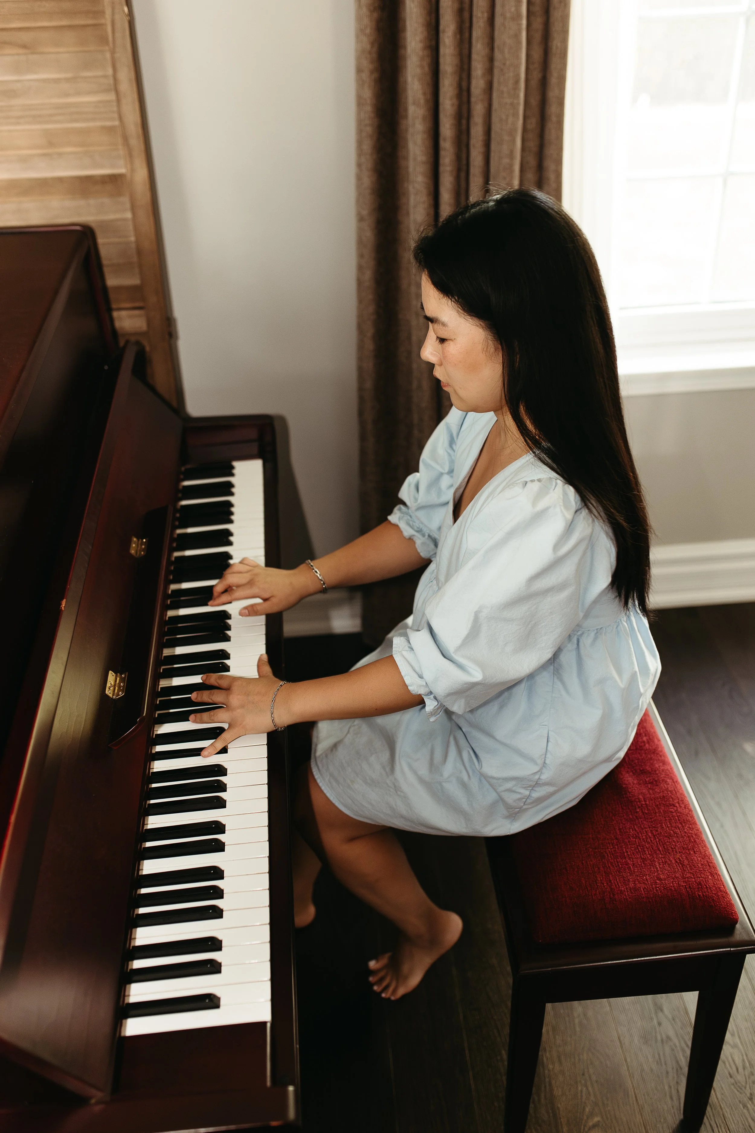 A woman with long dark hair wearing a light blue dress, sitting on a red cushioned bench, playing a wooden upright piano in a room with natural light coming through a window with beige curtains.