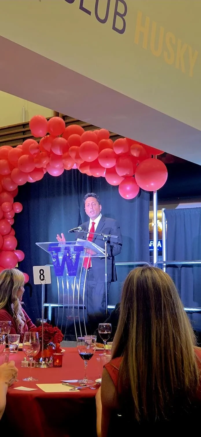 Ravi Vedula speaking at a podium with a University of Washington “W” logo during the Alpha Phi Red Dress Gala at Husky Stadium. He is dressed in a dark suit with a red tie, standing beneath a red balloon arch while addressing the audience on stage.