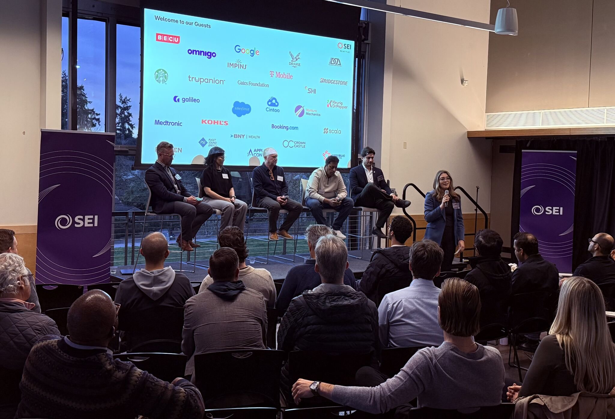 Crowd watching four panelists on stage beneath a large purple screen reading “SEI – AI Panel & Networking Event: From Hype to ROI"