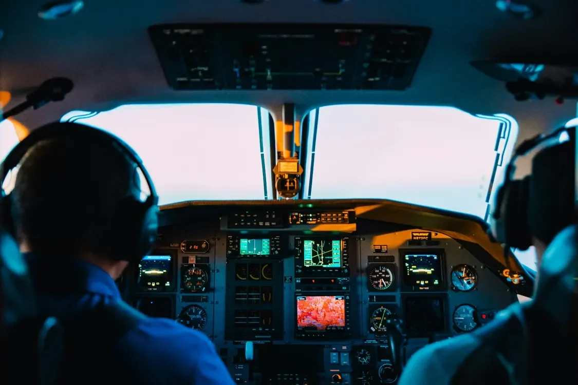 Two pilots wearing headsets sit in a cockpit, facing illuminated flight instruments and a bright sky through the windshield.
