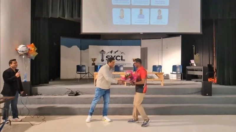 A person on stage presenting a bouquet to another speaker during an auditorium event, while a host stands nearby holding a microphone.