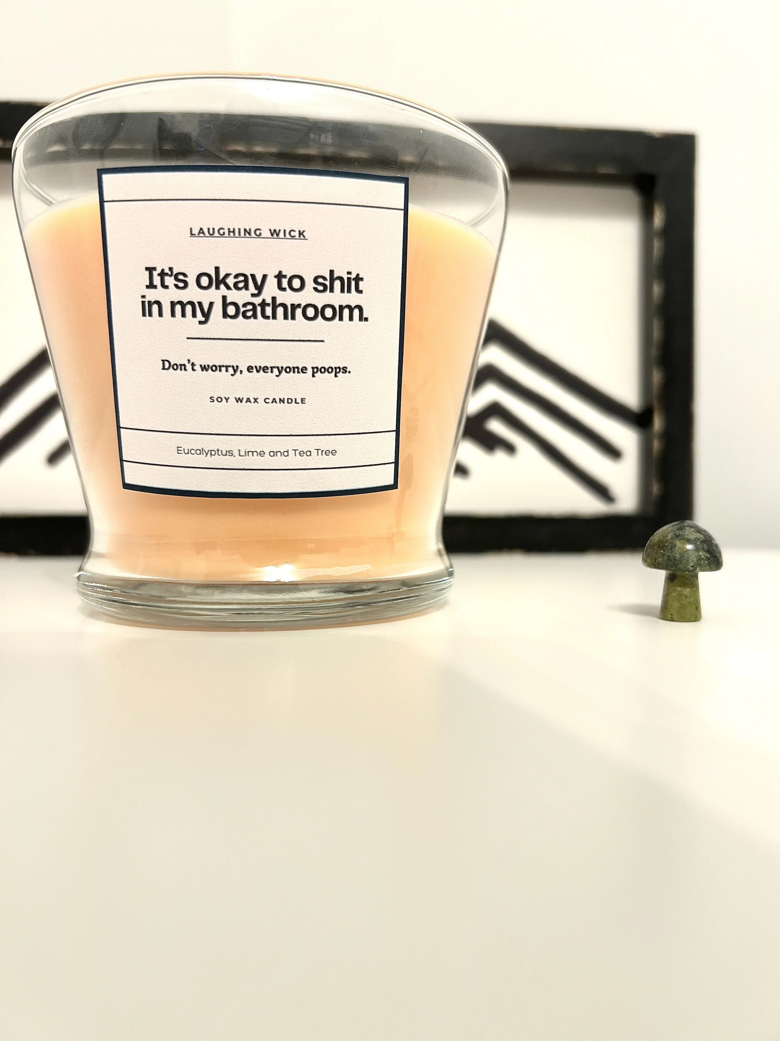 A pink soy wax candle in a clear glass container with a label that reads: "Laughing Wick: It's okay to shit in my bathroom. Don't worry, everyone poops. Soy wax candle. Eucalyptus, Lime and Tea Tree." Small decorative mushroom figurine on white surface in front of a black and white abstract art piece in the background.