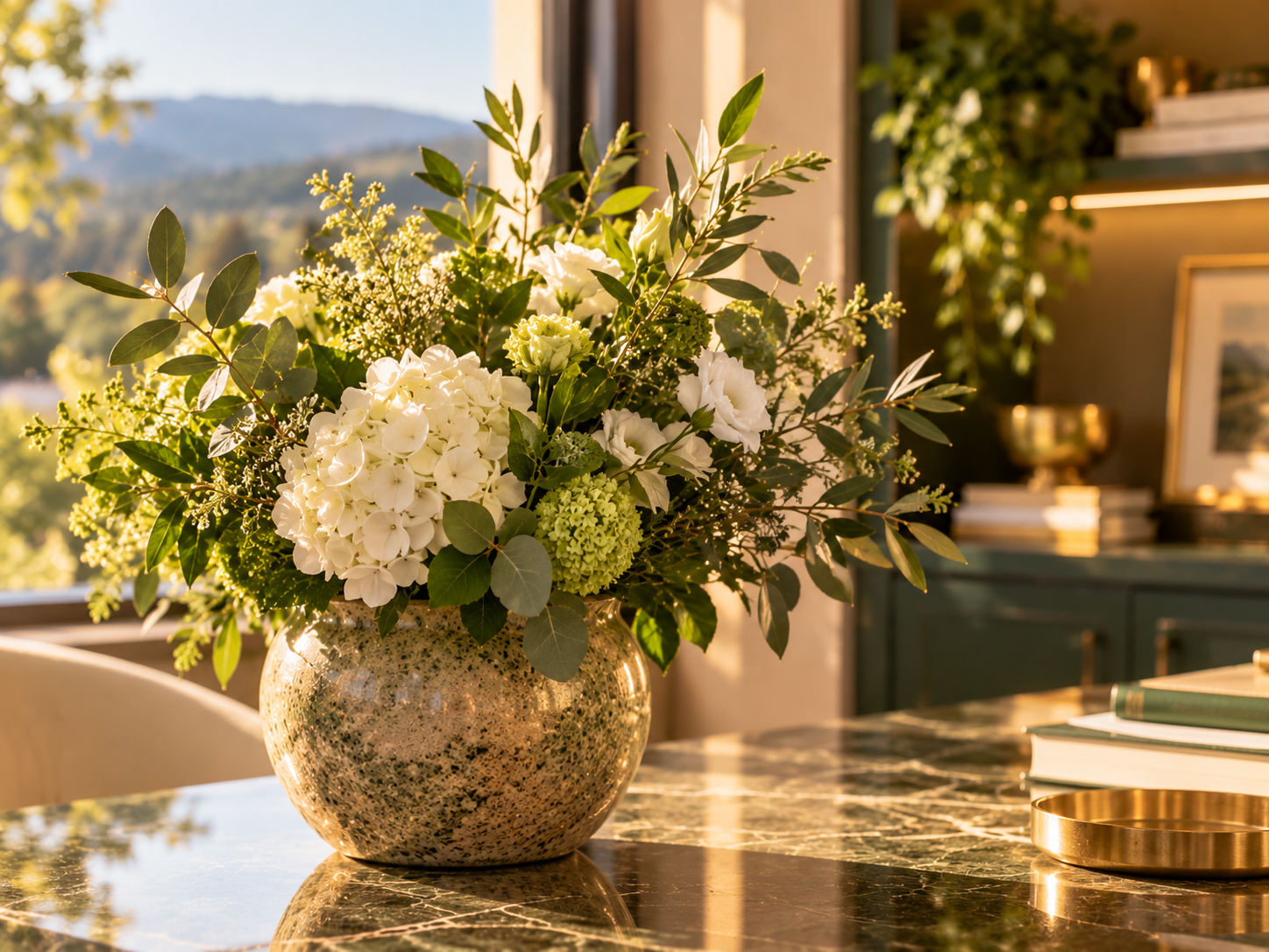 A floral arrangement of white flowers and greenery in a textured ceramic vase on a marble table, with sunlight streaming in and a window view of mountains in the background.
