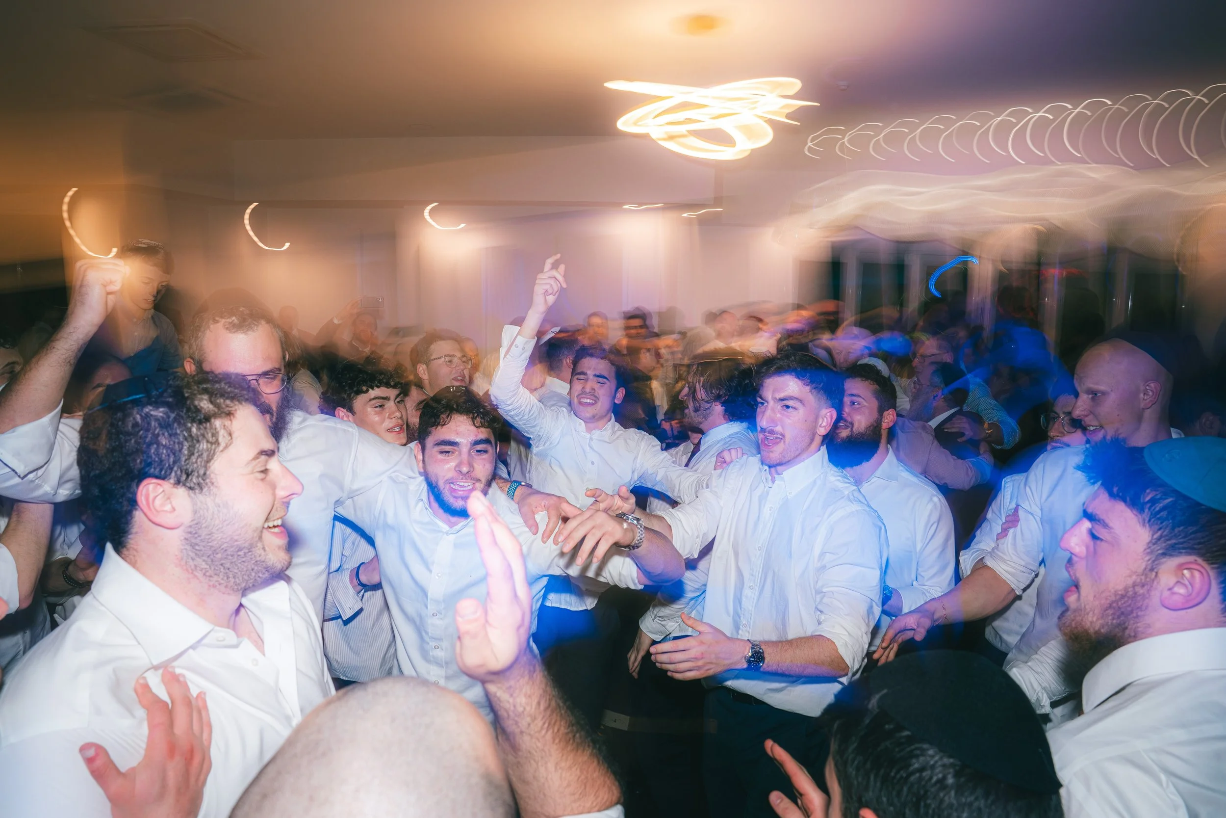 Group of young men dancing and celebrating at a party or wedding reception indoors with colorful motion-blurred lights.