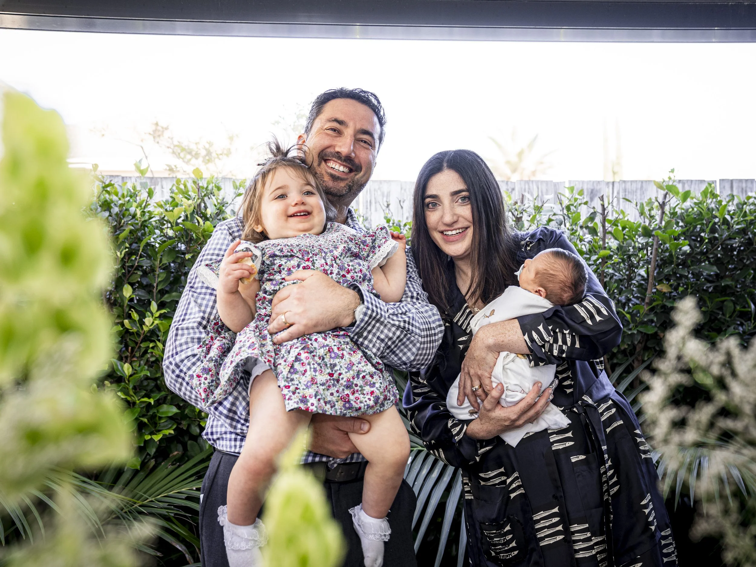 A happy family of four outdoors, with a man holding a young girl, and a woman holding a newborn baby, standing in front of greenery and a wooden fence.