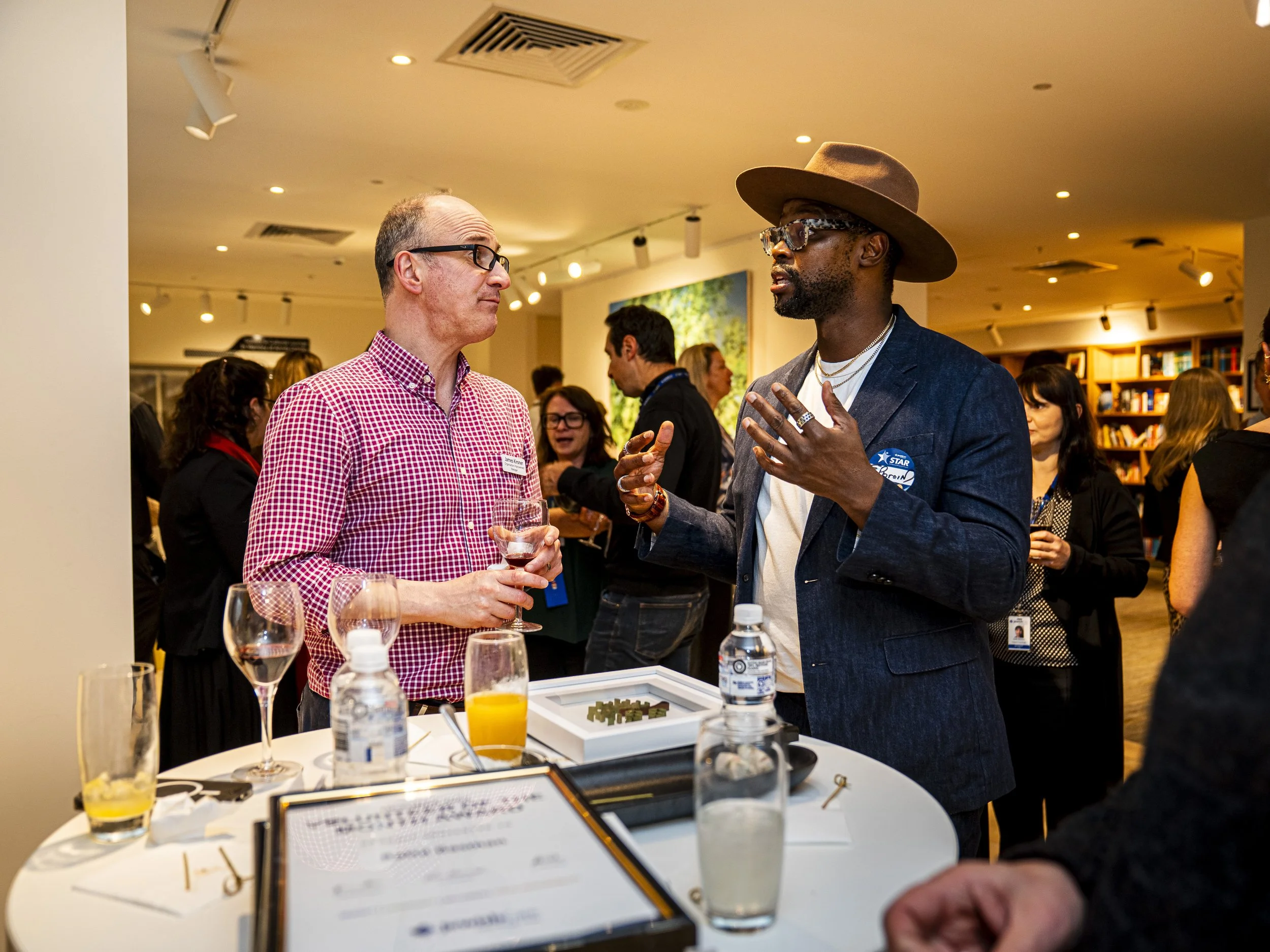 Two men engaged in conversation at a social event, one wearing a red checkered shirt holding a glass of red wine, the other wearing a dark blazer, hat, and glasses, gesturing with his hands, surrounded by people and drinks.