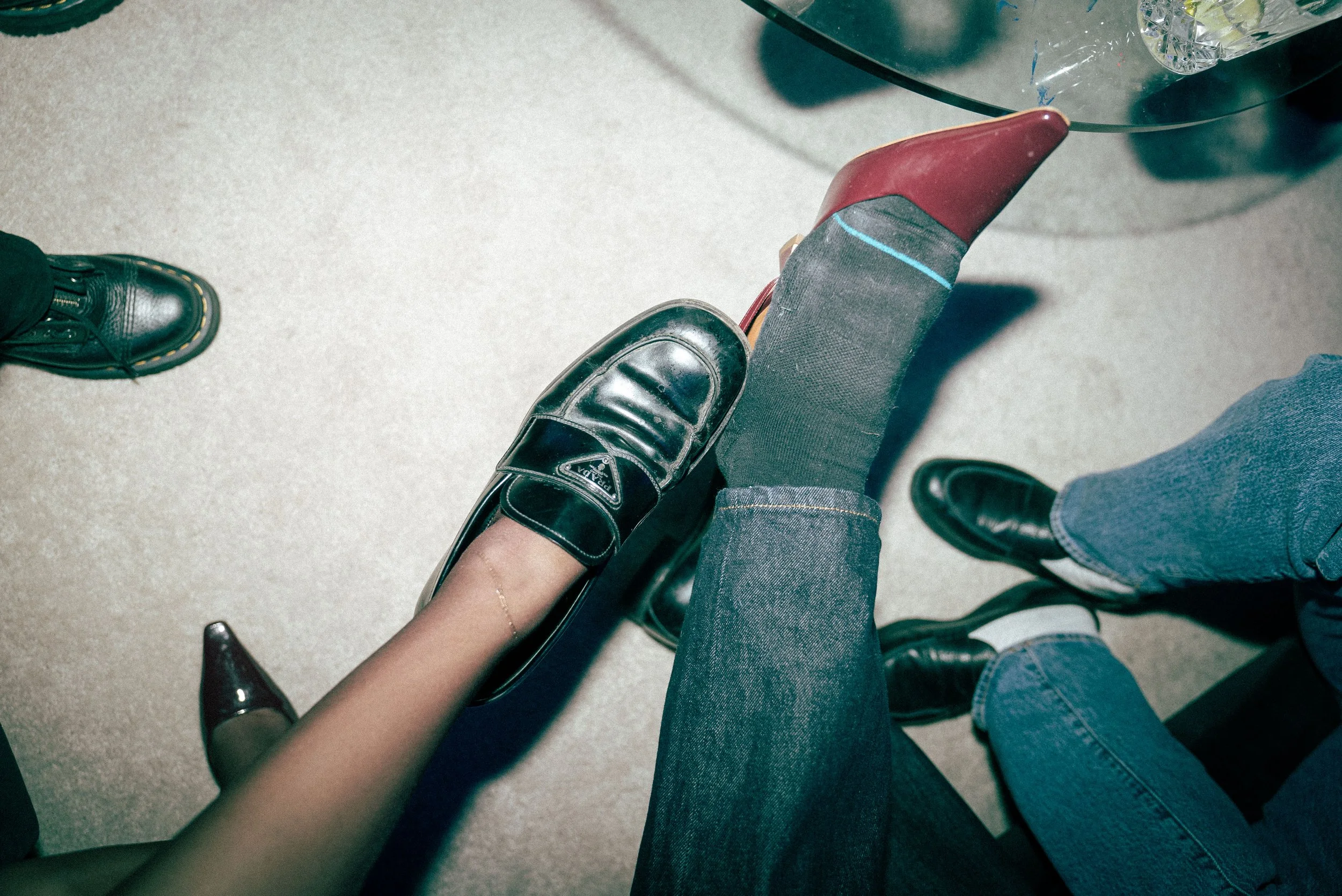 View from above of three people standing around, showing their feet and legs. The person in the center is sitting on a high-heeled shoe with one foot resting on a red shoe and the other foot on the ground. The other people are wearing black boots and jeans.