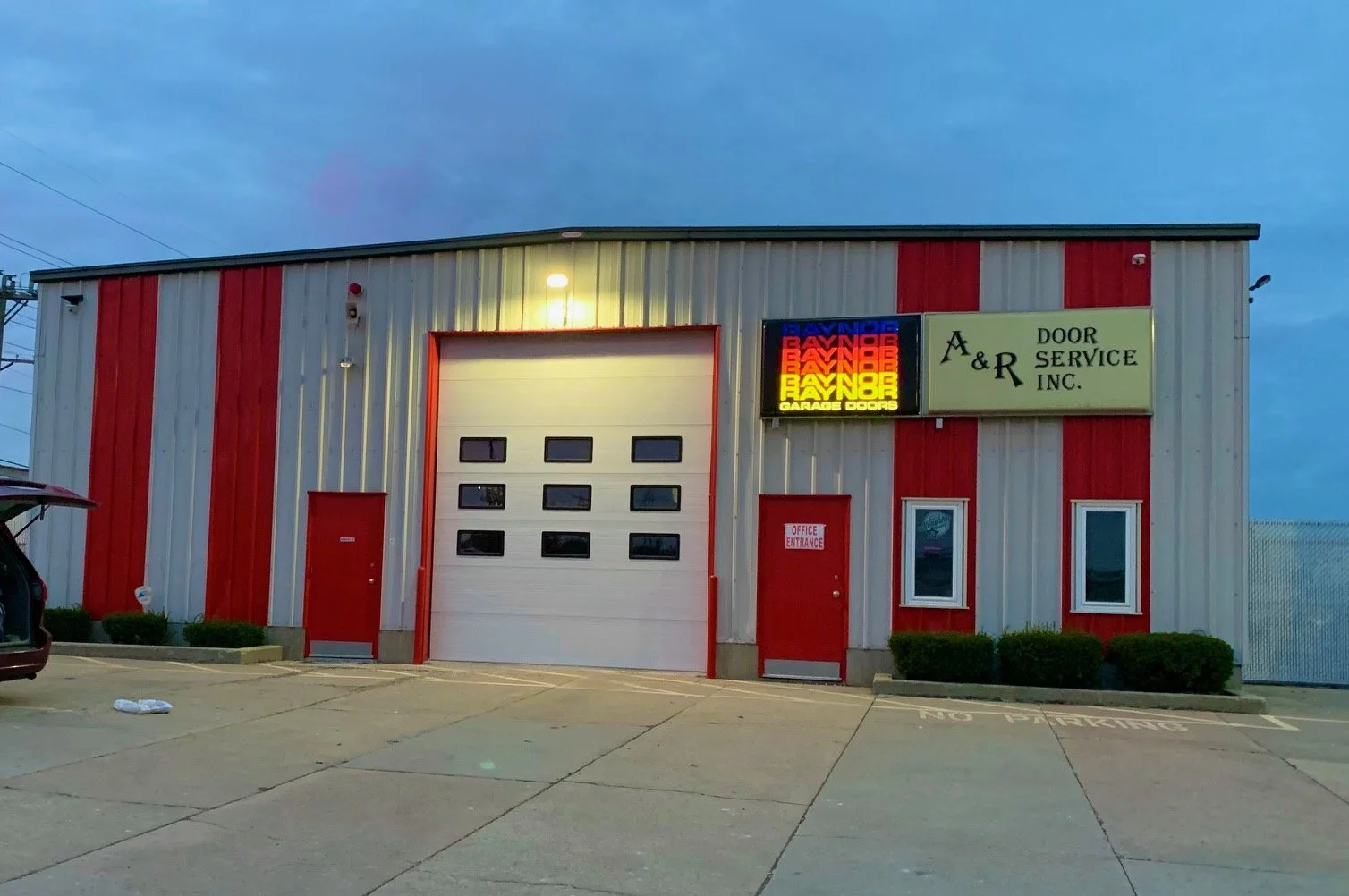 A commercial building with a large white garage door and red doors, two small windows, bushes, and a neon sign that reads "Baynor Garage Doors" and "A & R Door Service Inc." in a parking lot at dusk.