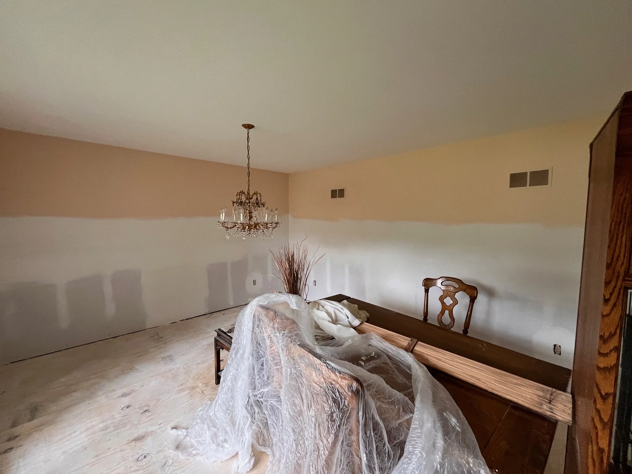 Partially painted living room with beige and white walls, a chandelier hanging from the ceiling, a wooden table, a chair, and a plastic sheet draped over furniture.