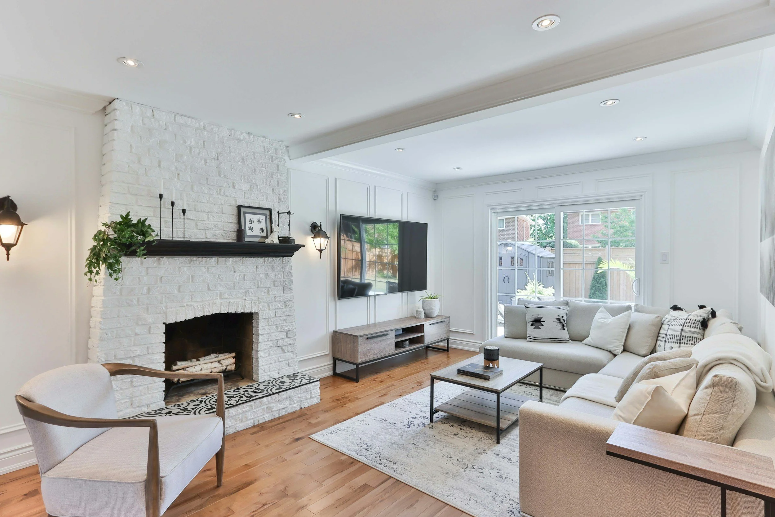 Living room with white brick fireplace, flat-screen TV on a wooden stand, beige L-shaped sofa, white armchair, wooden coffee table, large window, hardwood floors, and decorative wall lights.