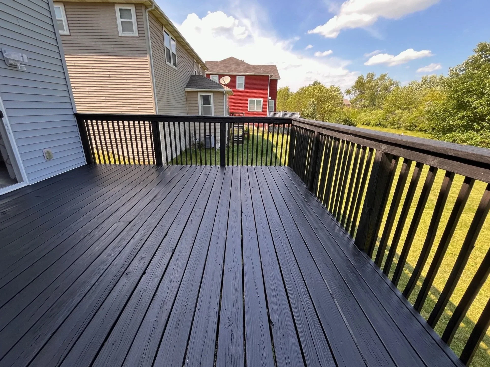 Empty wooden balcony with black railings overlooking neighboring houses and a grassy backyard, under a partly cloudy blue sky.