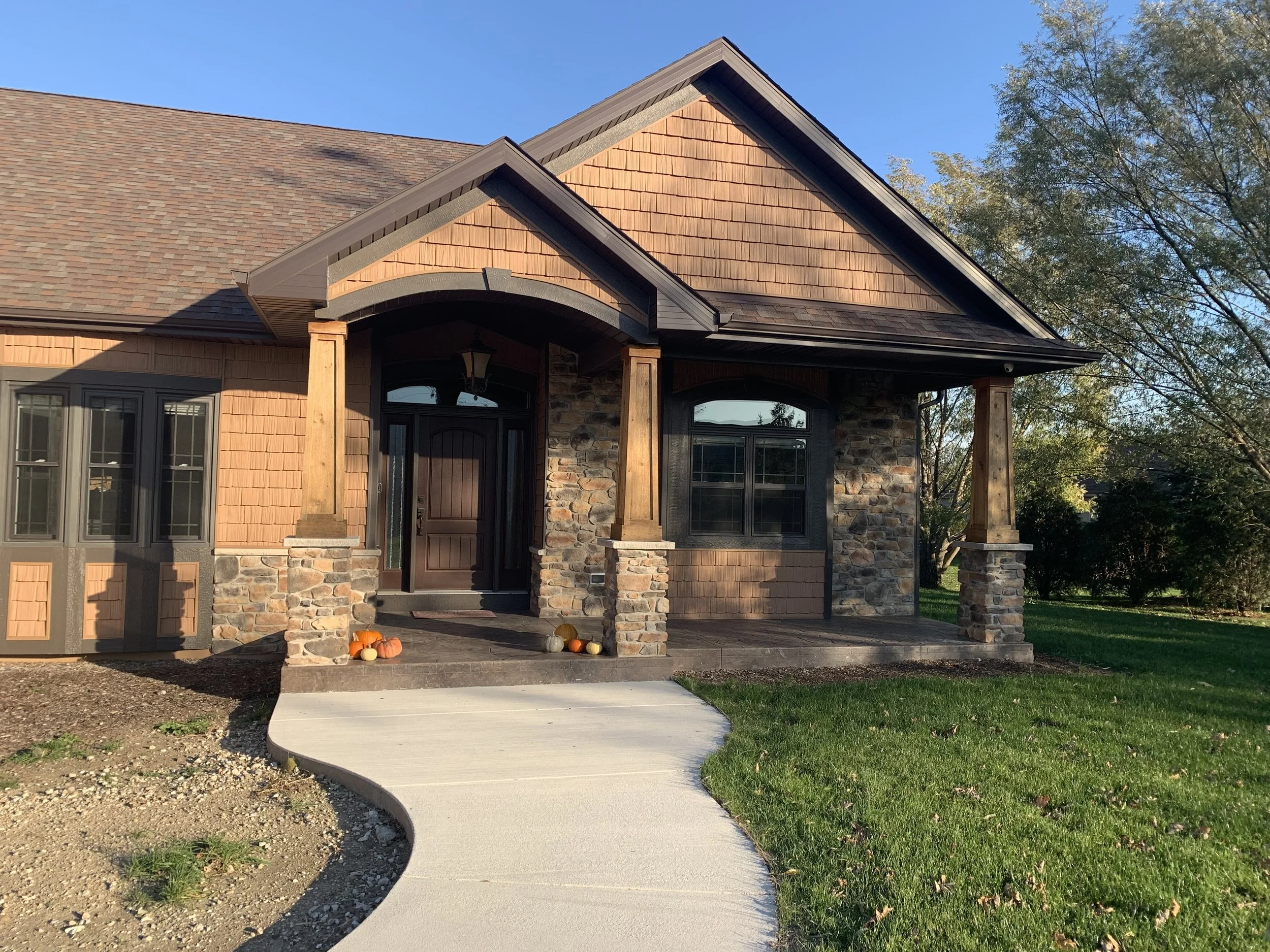 Front porch of a house with stone and wood exterior, arched doorway, a small porch with pumpkins, and a curved concrete walkway surrounded by grass and trees.
