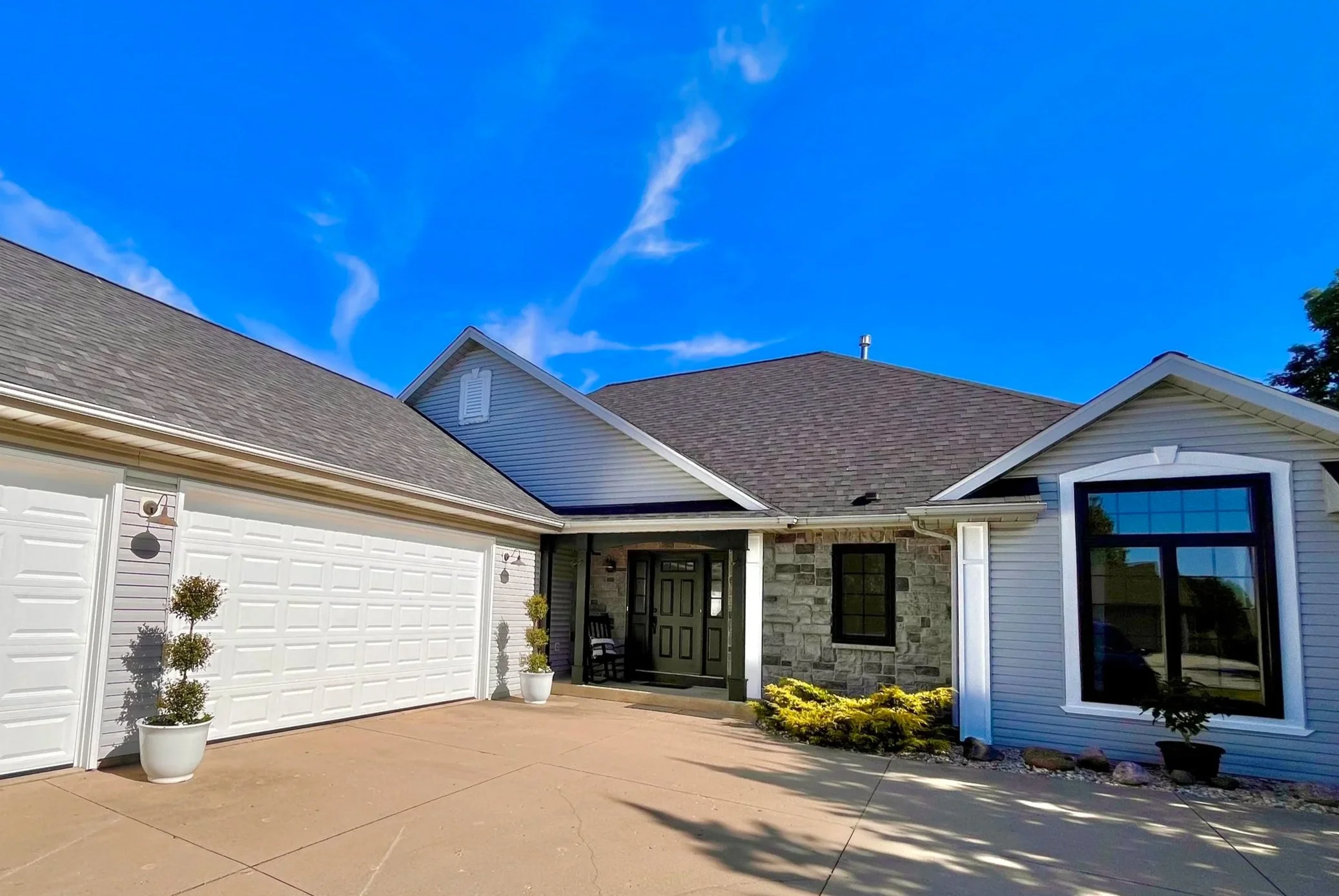 Front view of a house with a driveway, white garage door, black front door, large window, and landscaped front yard against a bright blue sky with some clouds.