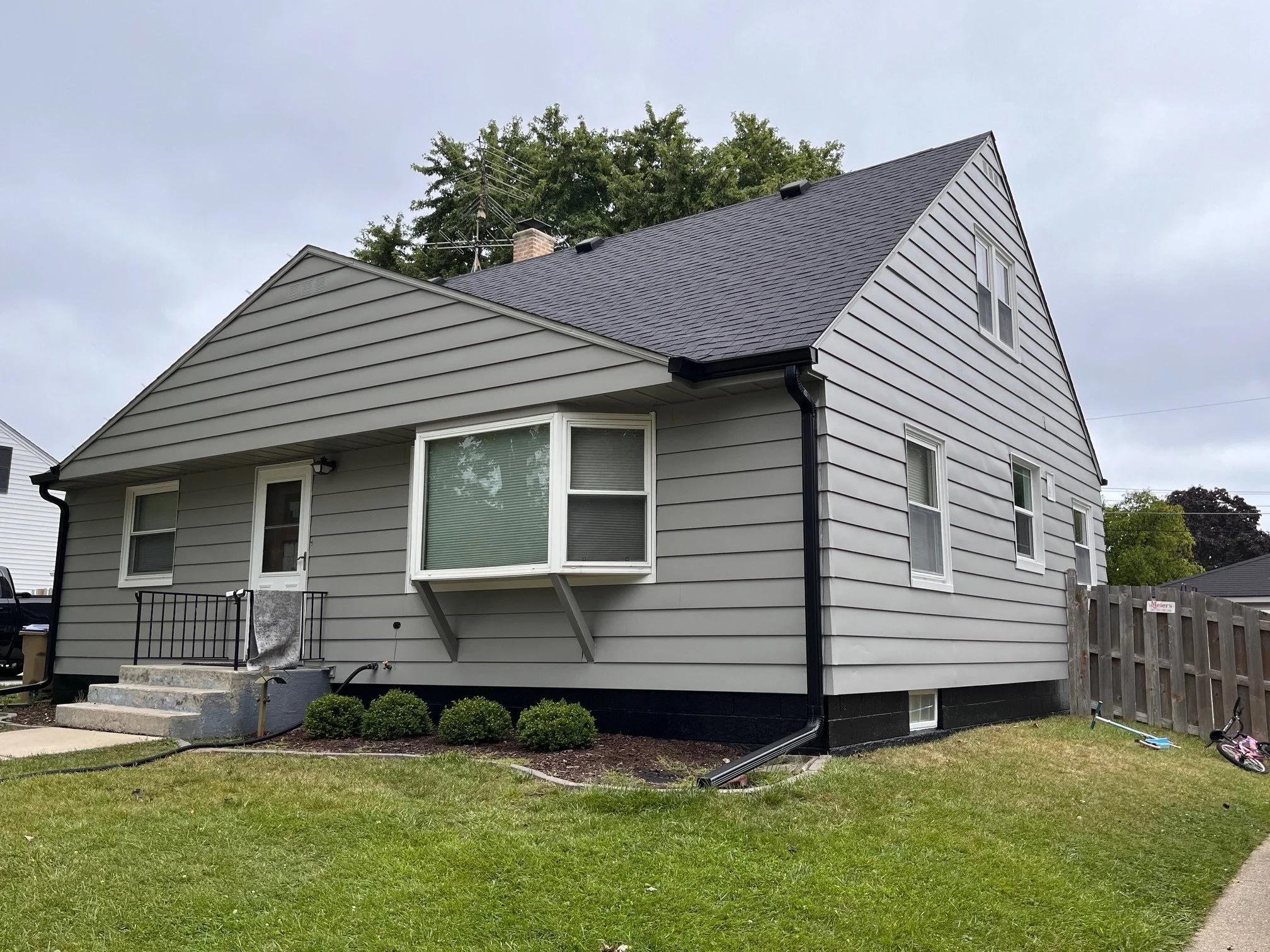 Gray two-story house with a small porch, black railing, and steps, surrounded by a green lawn with bushes and a fence, under a cloudy sky.