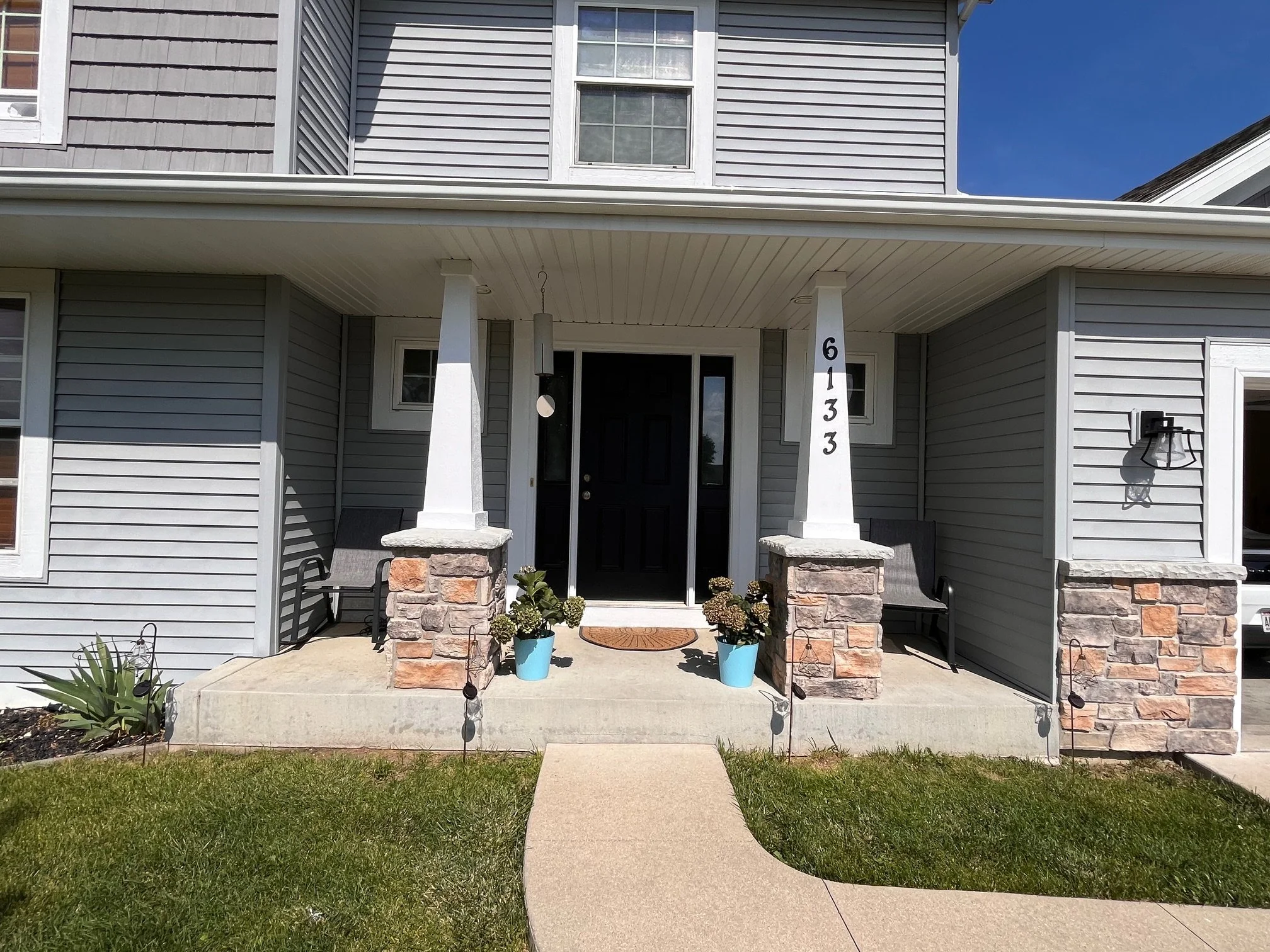 Front porch of a house with gray siding, black front door, two chairs, potted plants, and house number 6133, under a porch roof with a clear blue sky.