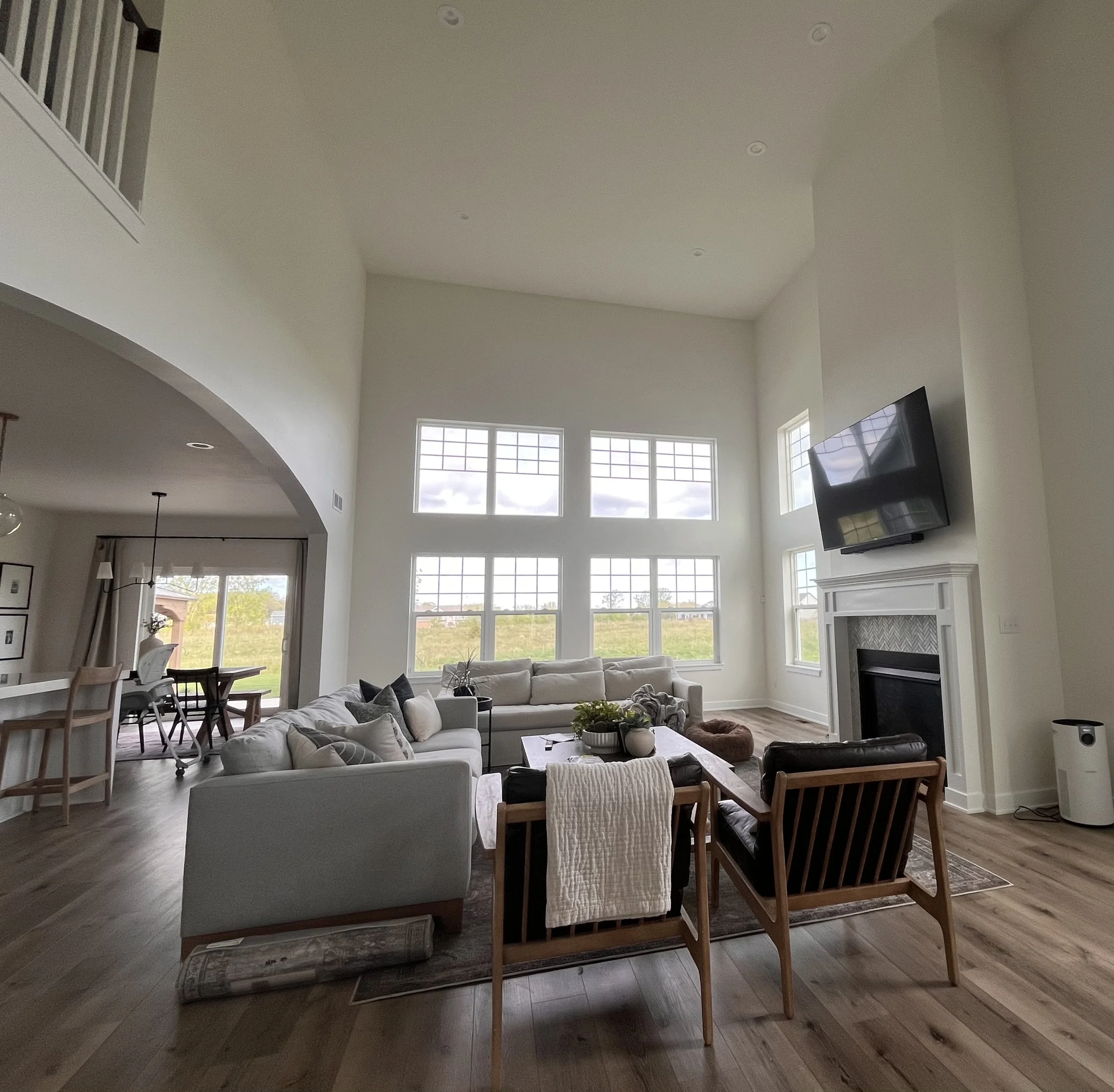 Bright living room with large windows, white couches, wooden armchairs, and a coffee table, with a fireplace and TV mounted above it.
