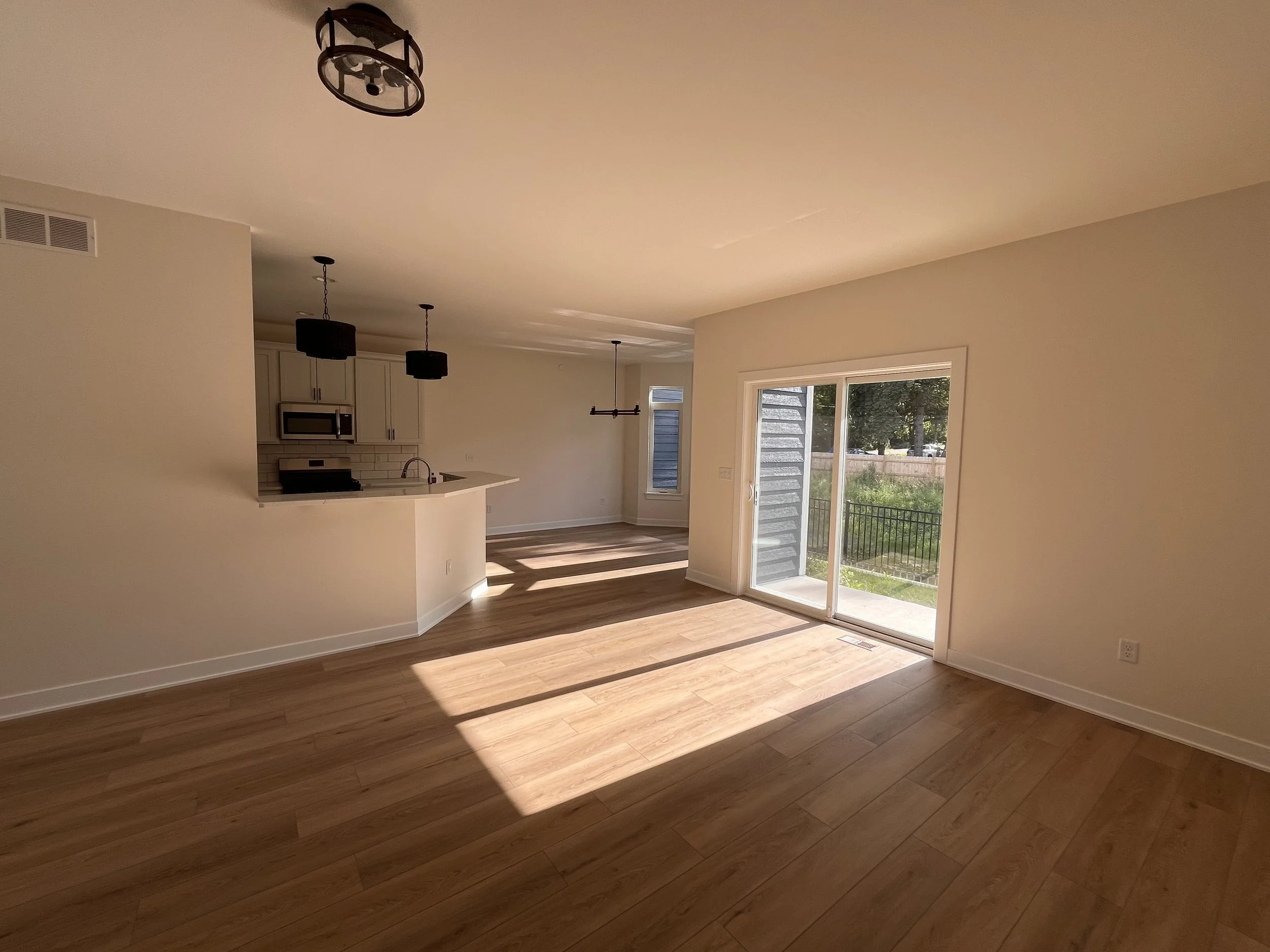 Empty living room with hardwood floors, large sliding glass door leading outside, and open kitchen in the background with white cabinetry, a microwave, and pendant lighting.