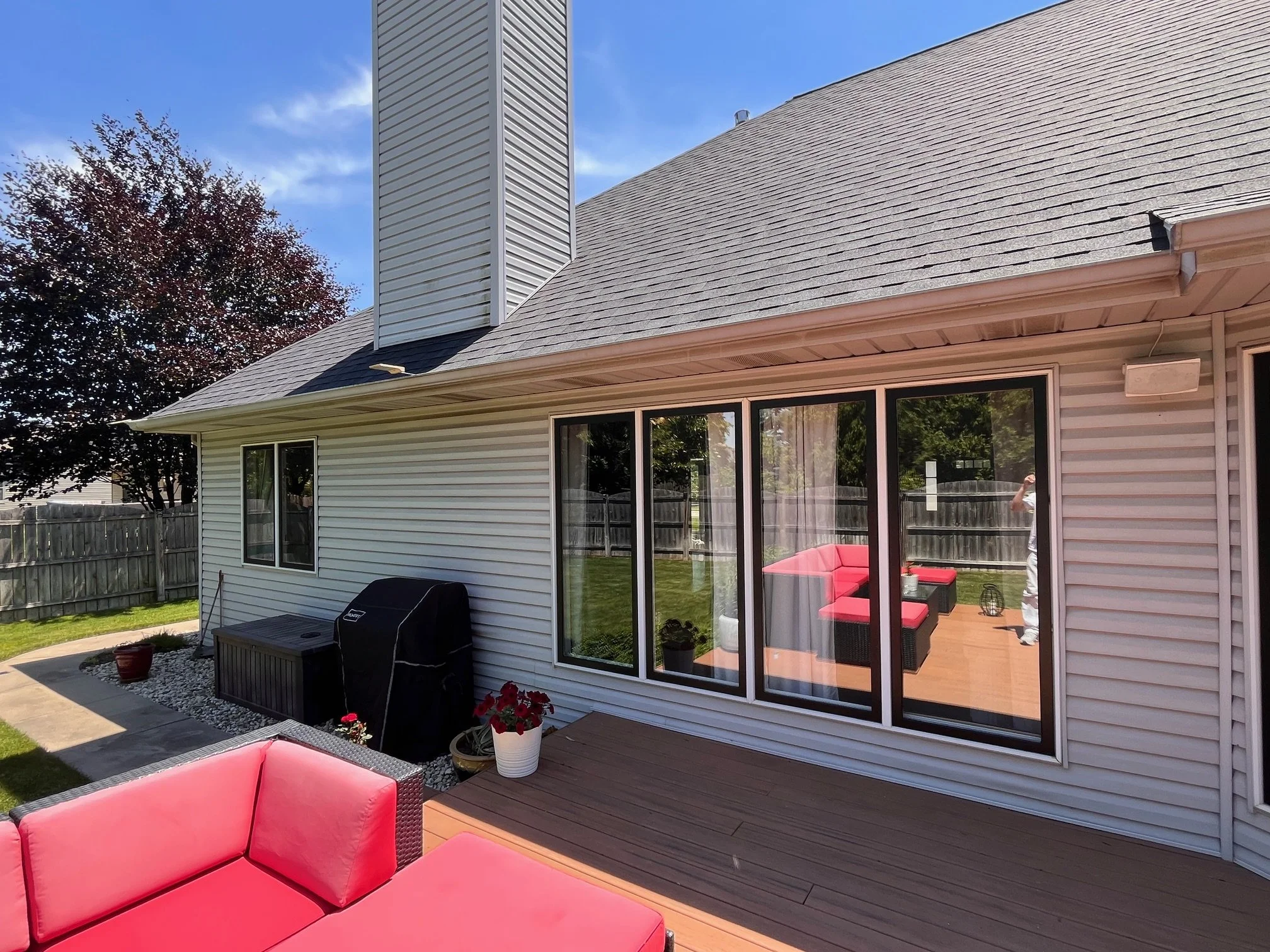 Backyard patio with pink outdoor furniture, a barbecue grill, potted plants, a wooden deck, and a sliding glass door leading into a house.
