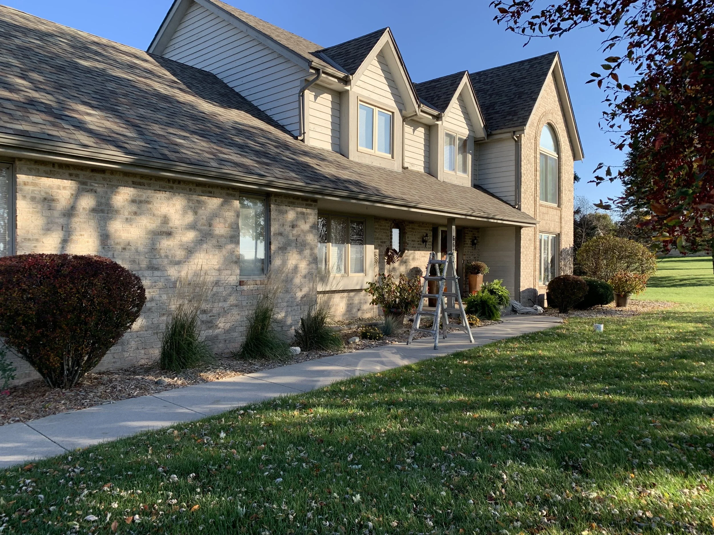 A two-story house with brick and siding exterior, multiple windows, and a front porch with potted plants. A ladder is set up on the walkway, and the yard has grass, shrubs, and trees. It appears to be late afternoon or early evening.