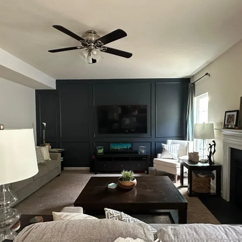 Living room with a black accent wall, white walls, ceiling fan, large flat-screen TV, beige sofa, white armchair, side tables with lamps, fireplace, and a window with curtains.