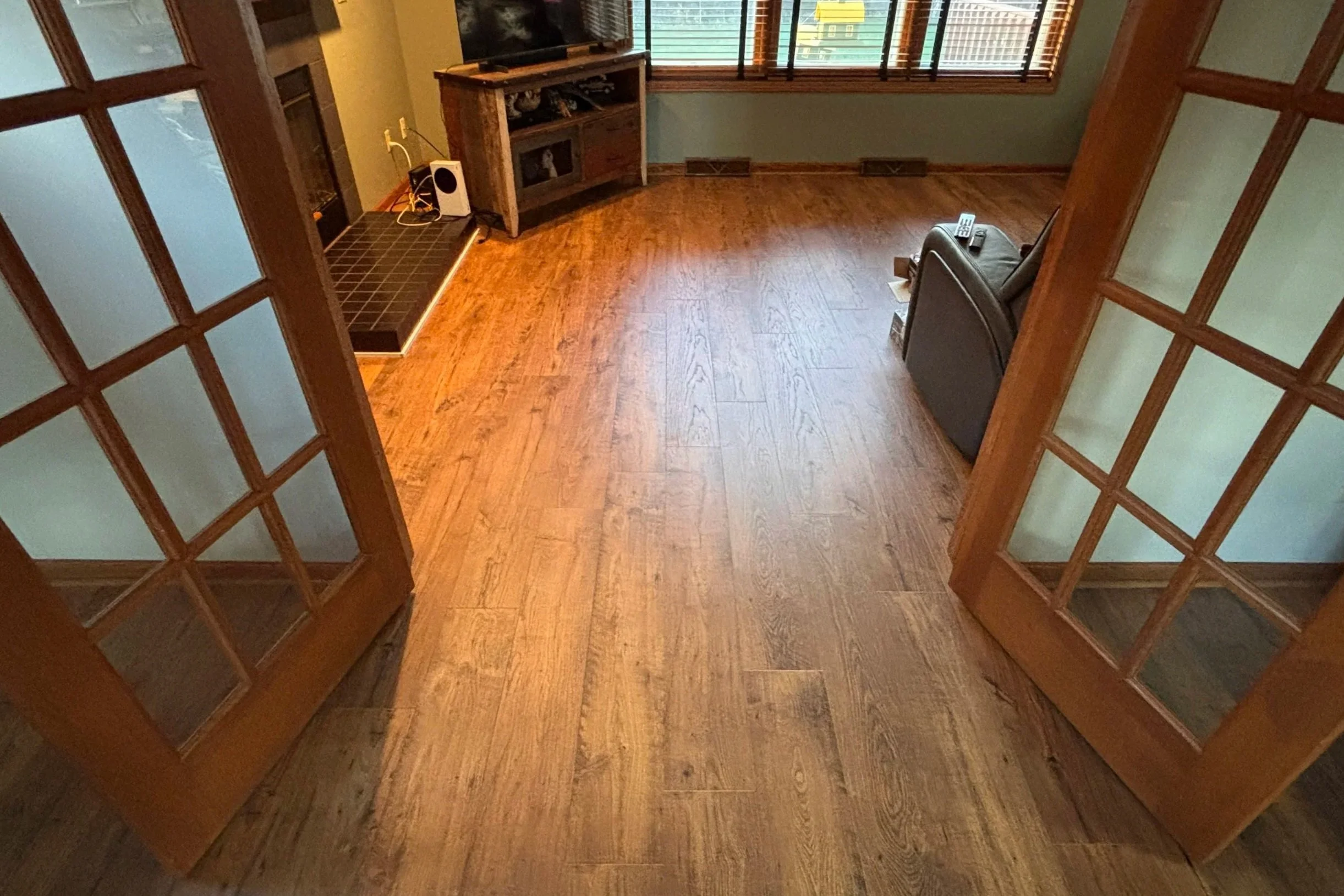 Living room with wooden floor, glass-paneled double doors, a TV on a wooden stand, a chair with a remote, and windows with blinds.