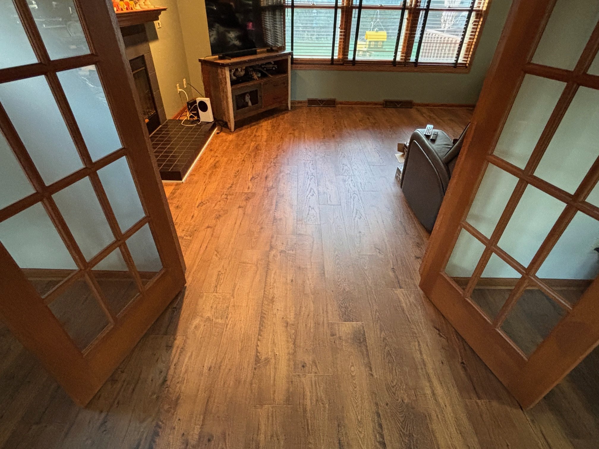View of a living room with wooden floors, large windows with blinds, a TV on a wooden stand, a black armchair, a remote control, and a small black tiled area near the corner.
