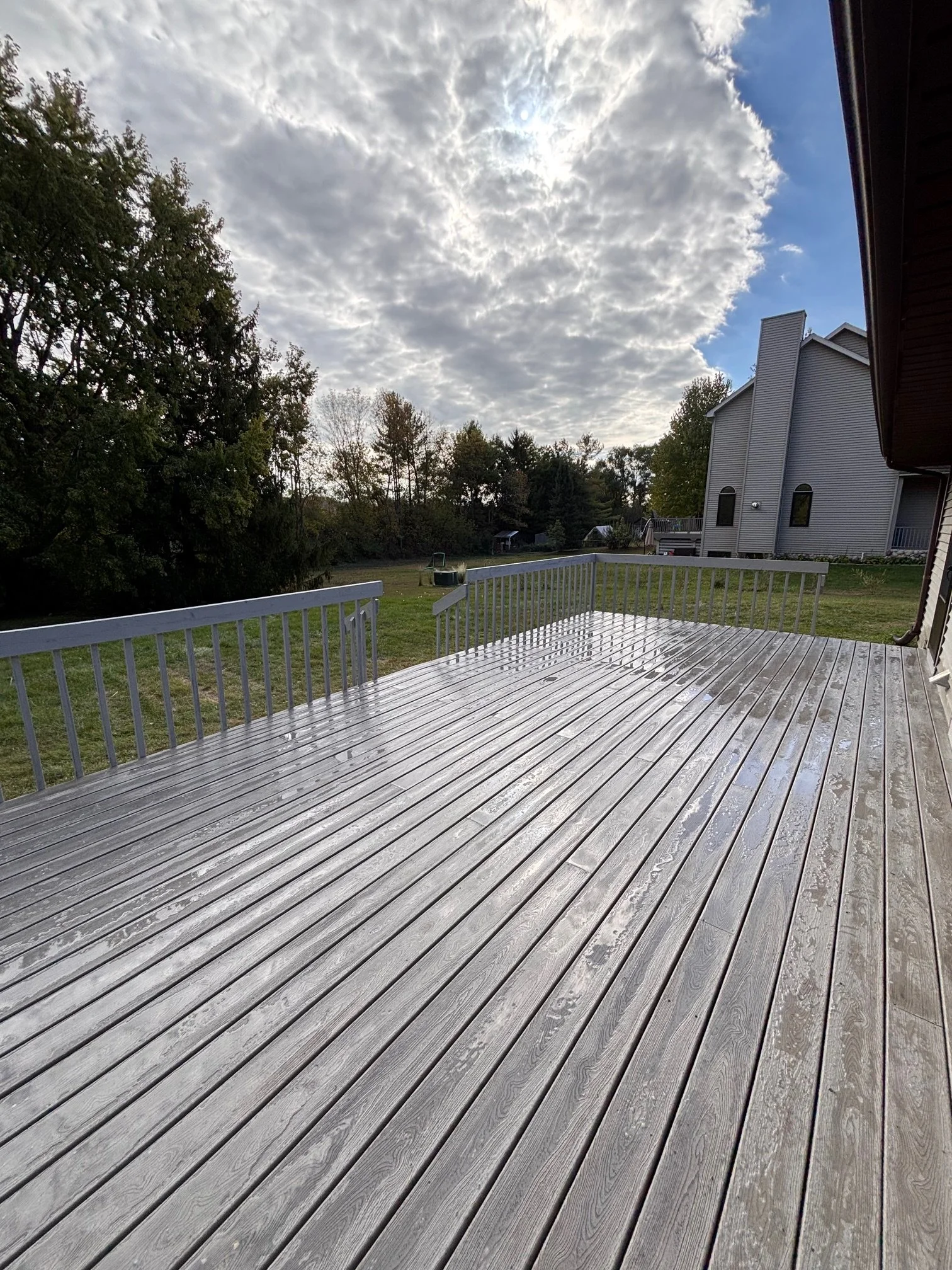 Clean, wet wooden deck with a railing, overlooking a grassy yard with trees and a modern house, under a partly cloudy sky with the sun peeking through.