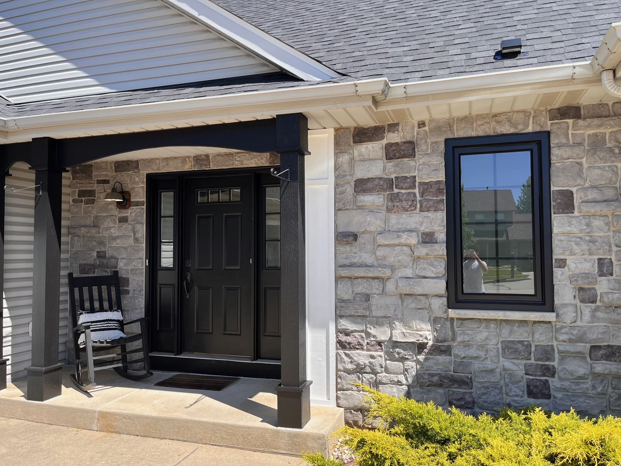 Front porch of a house with a black door, two small side windows, a rocking chair with a cushion, and a stone facade with a window reflecting the neighborhood.