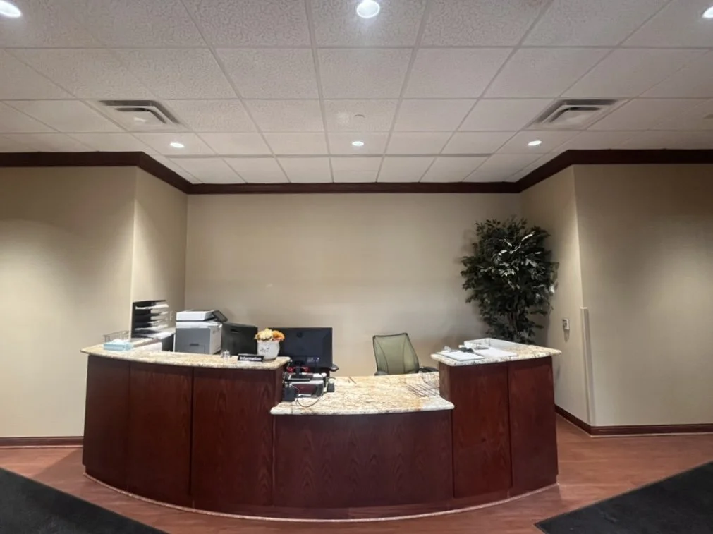 Empty reception desk with a computer, printer, and office supplies, located in a lobby area with beige walls, a tree in the corner, and wooden flooring.