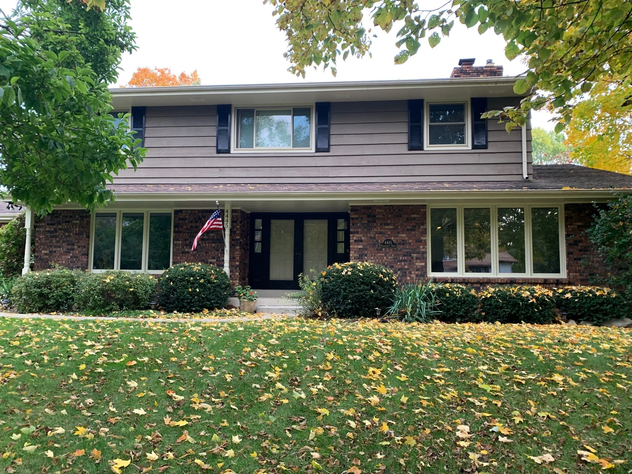 A two-story house with brick and gray siding exterior, front yard with fallen leaves, bushes, and trees, American flag near the front door, and large windows.
