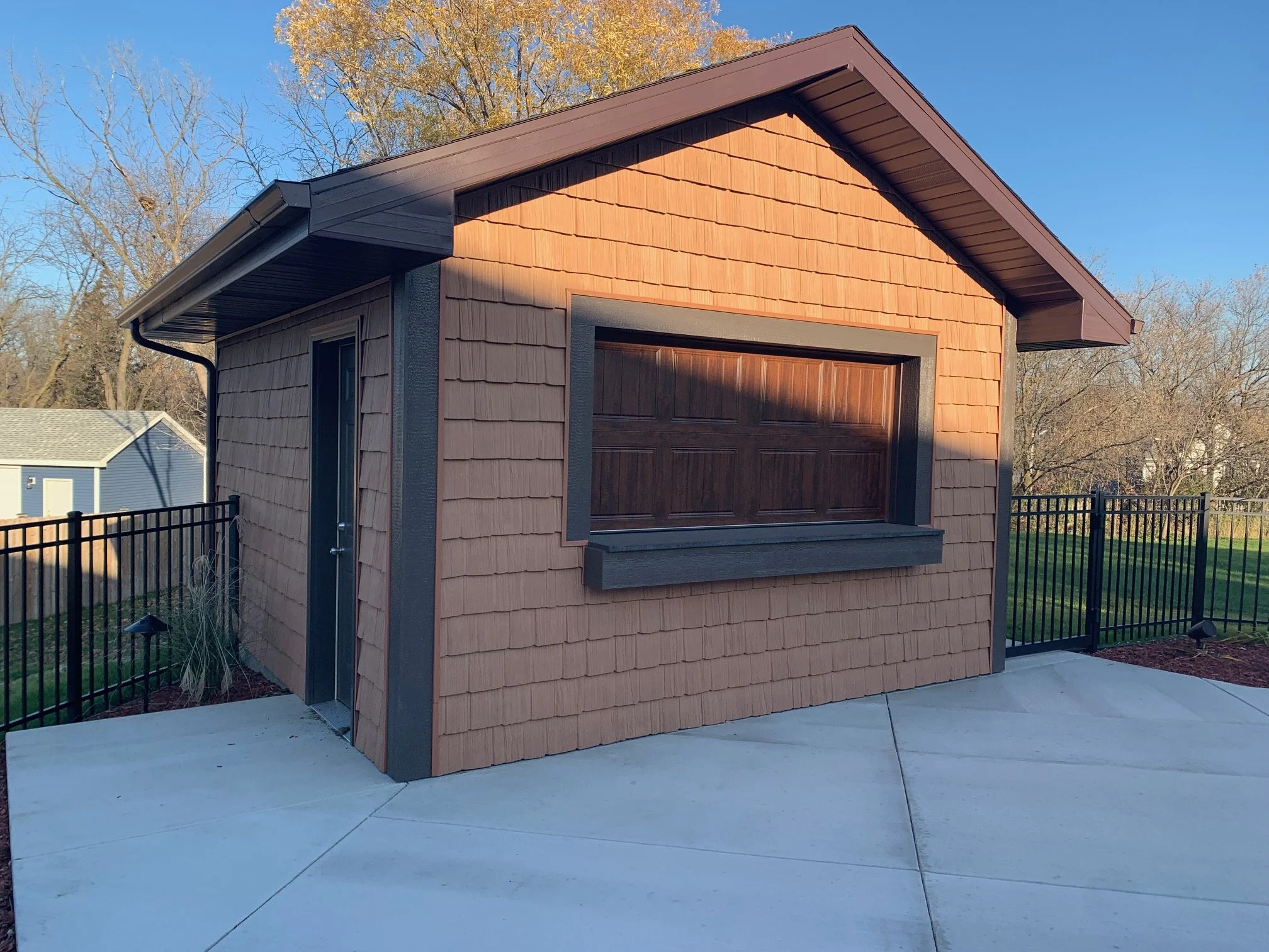 A small, wooden shed with a brown shingled exterior, dark brown window, and a small door, surrounded by a black metal fence, set in a backyard with trees and a concrete patio.