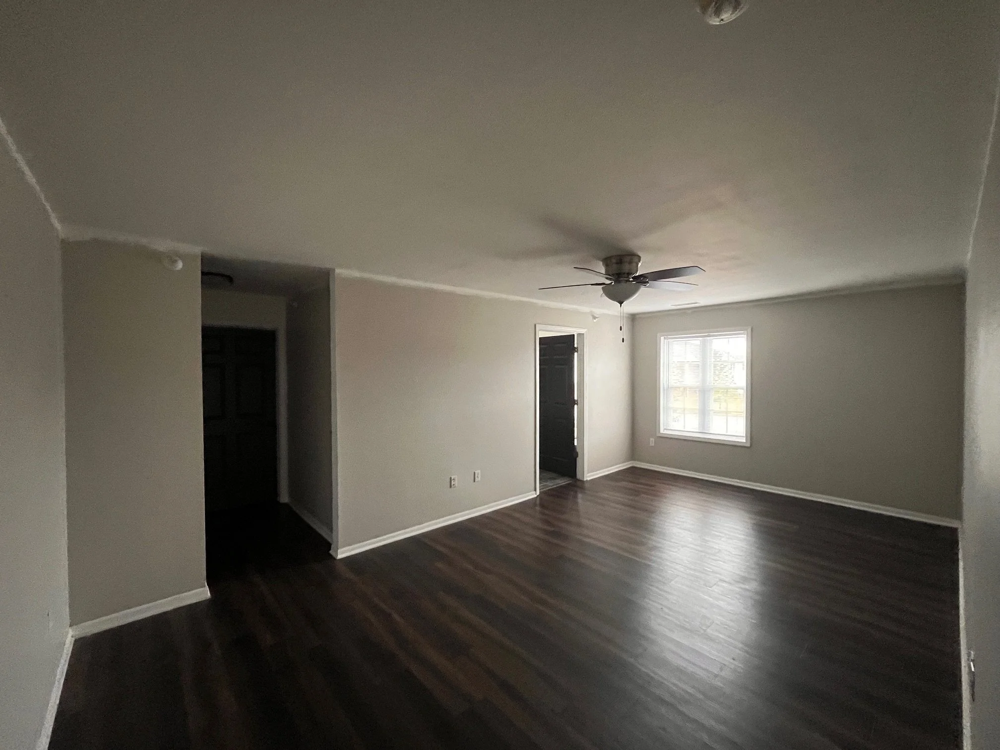 Empty living room with dark wood flooring, a ceiling fan, a window allowing natural light in, and an open door leading outside.