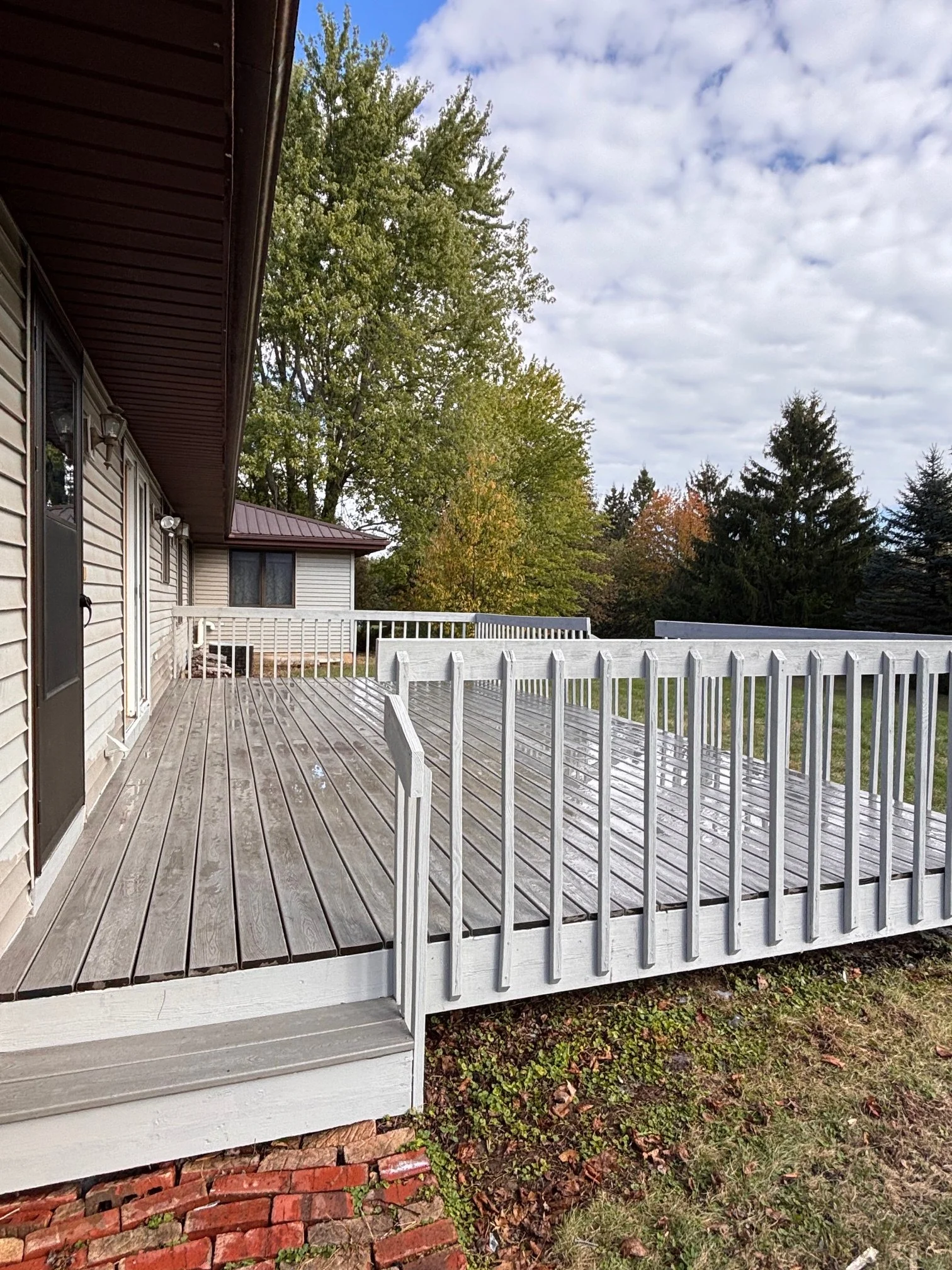 A wooden deck attached to a house with beige siding and a brown roof, surrounded by a white railing, with trees and a partly cloudy sky in the background.