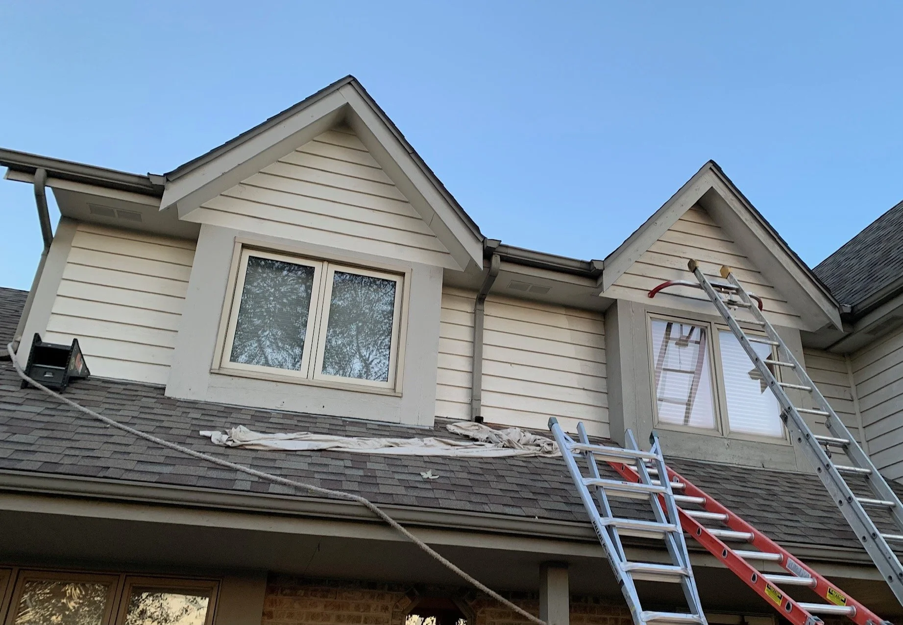 Ladders leaning against the roof of a house under maintenance or repair, with two large windows on the second floor and a pitched roof with shingles, under a clear blue sky.