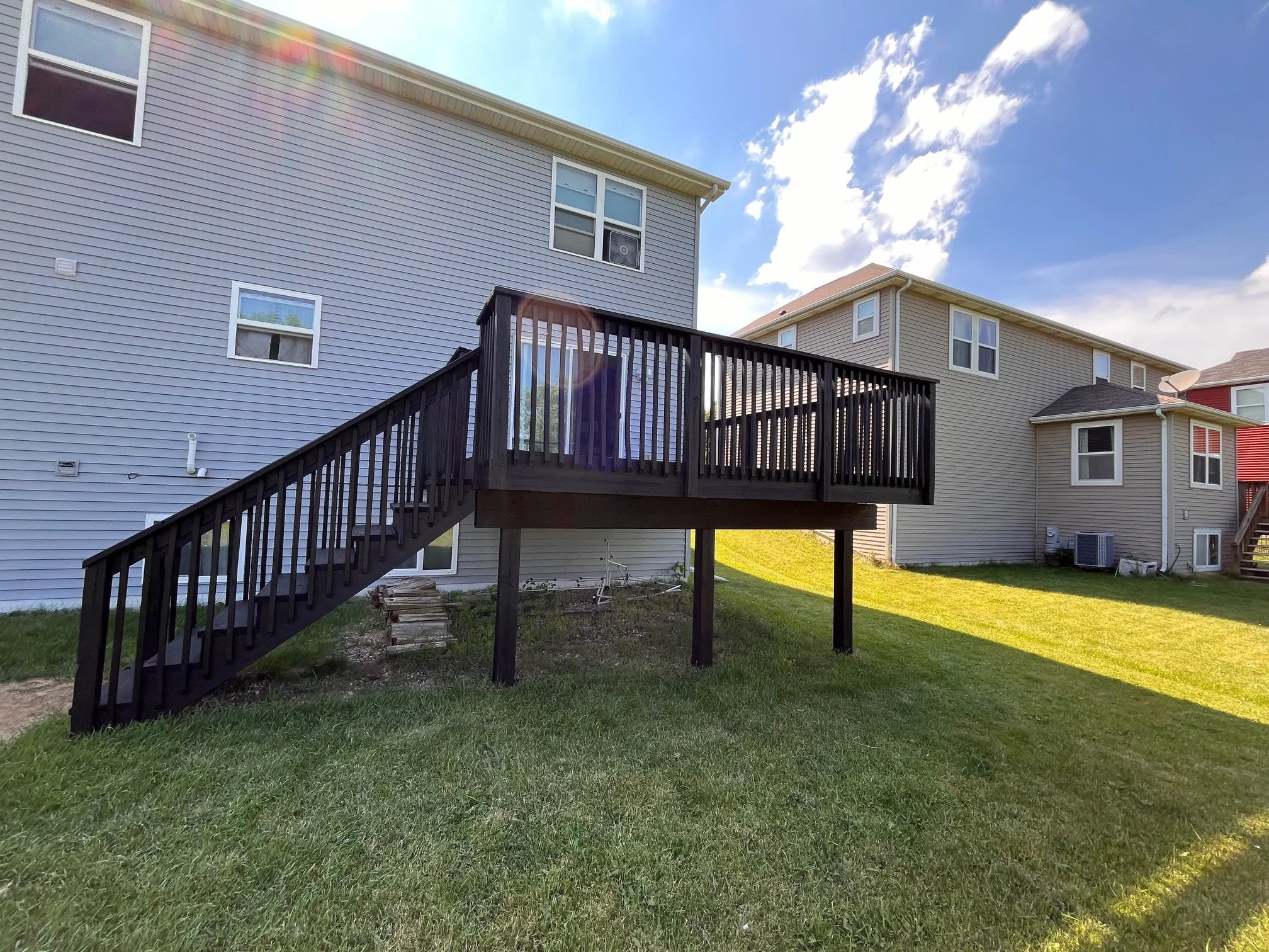 A backyard with a newly built wooden deck attached to a light blue house. The deck has stairs leading down to a grassy yard, and the sky is partly cloudy with sunshine.