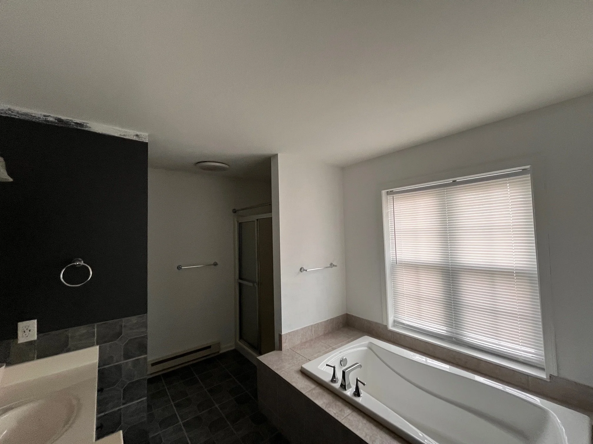 Bathroom with a large window with closed blinds, corner bathtub with faucets, and a dark tiled floor.