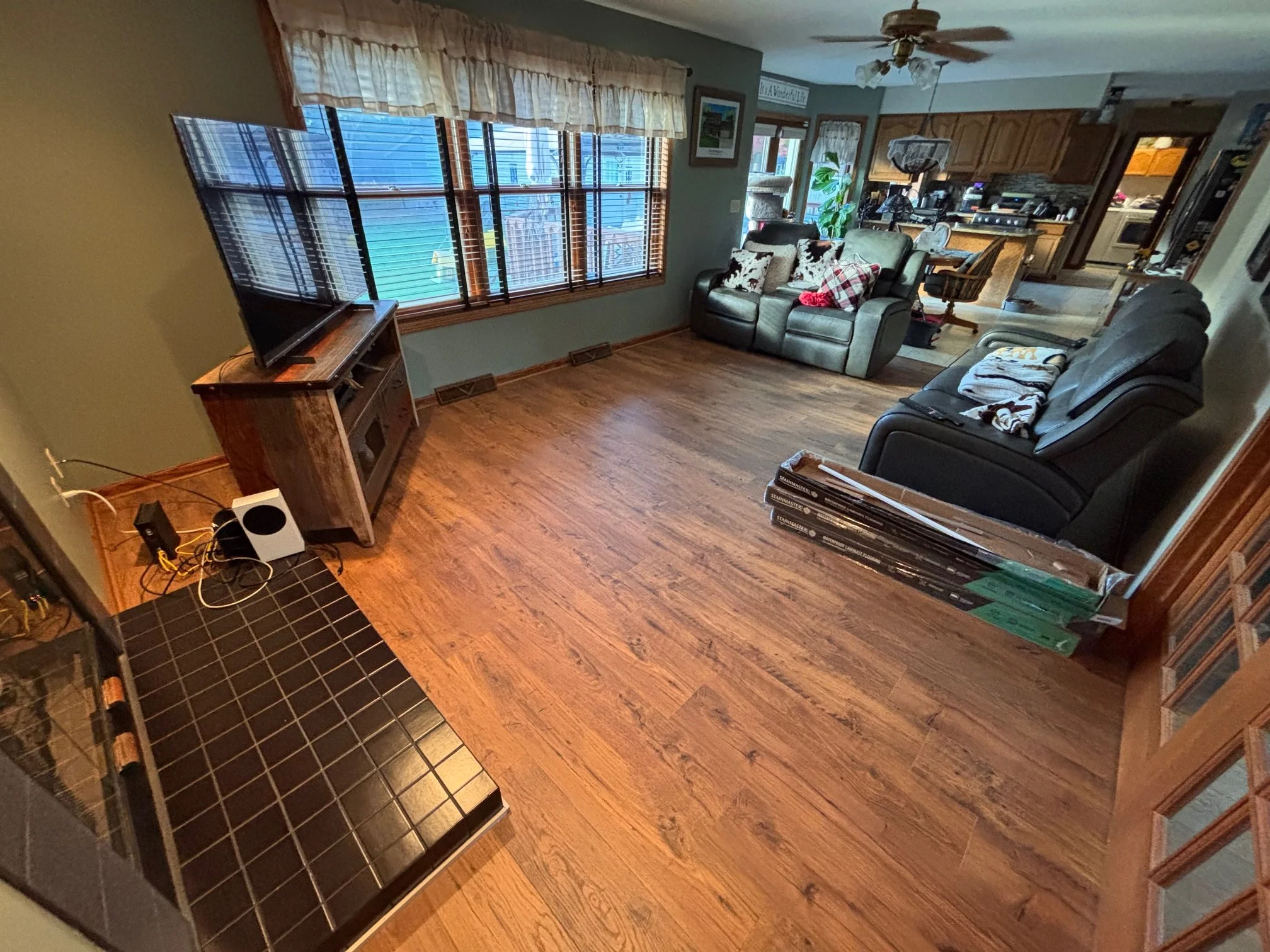 Living room with hardwood floors, green walls, a window with blinds, a flat-screen TV on a wooden stand, two black reclining sofas, a small section of black tiled hearth, and stacked flooring materials.