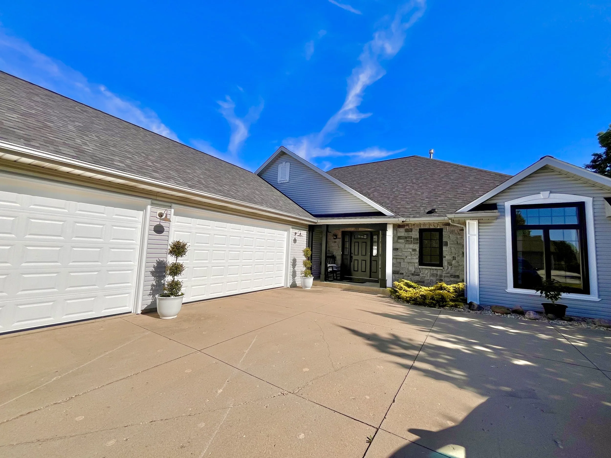 Front of a house with a driveway, garage doors, and a porch with a potted plant, blue sky with some clouds in the background.