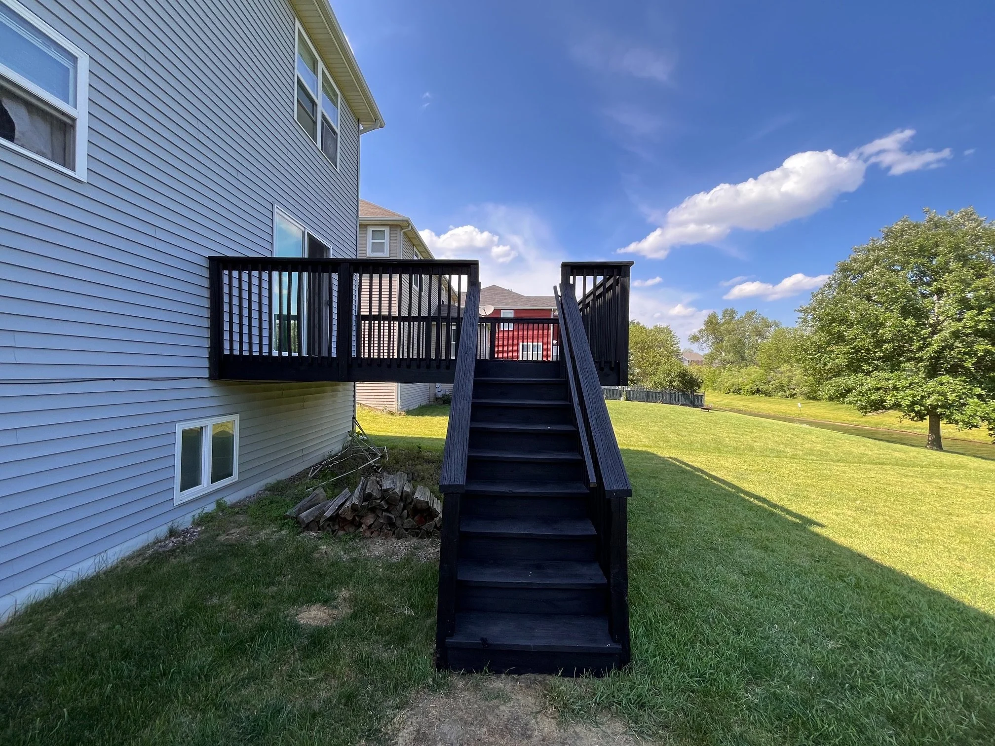 Backyard with a wooden staircase leading up to a small deck attached to a house with blue siding. The yard is green with some trees and a partly cloudy blue sky.