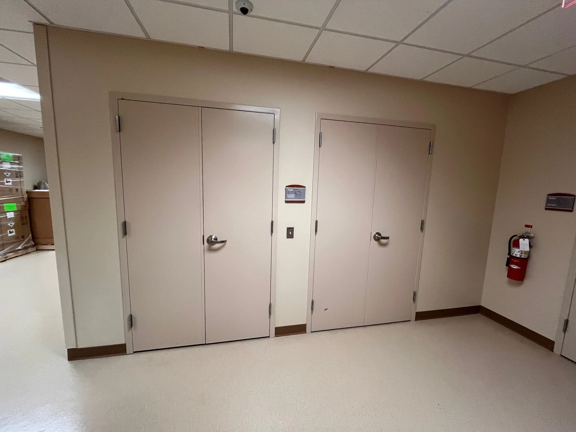 Two white doors in a beige wall inside a building, with a fire extinguisher mounted on the right wall and a small sign on the wall next to the doors.