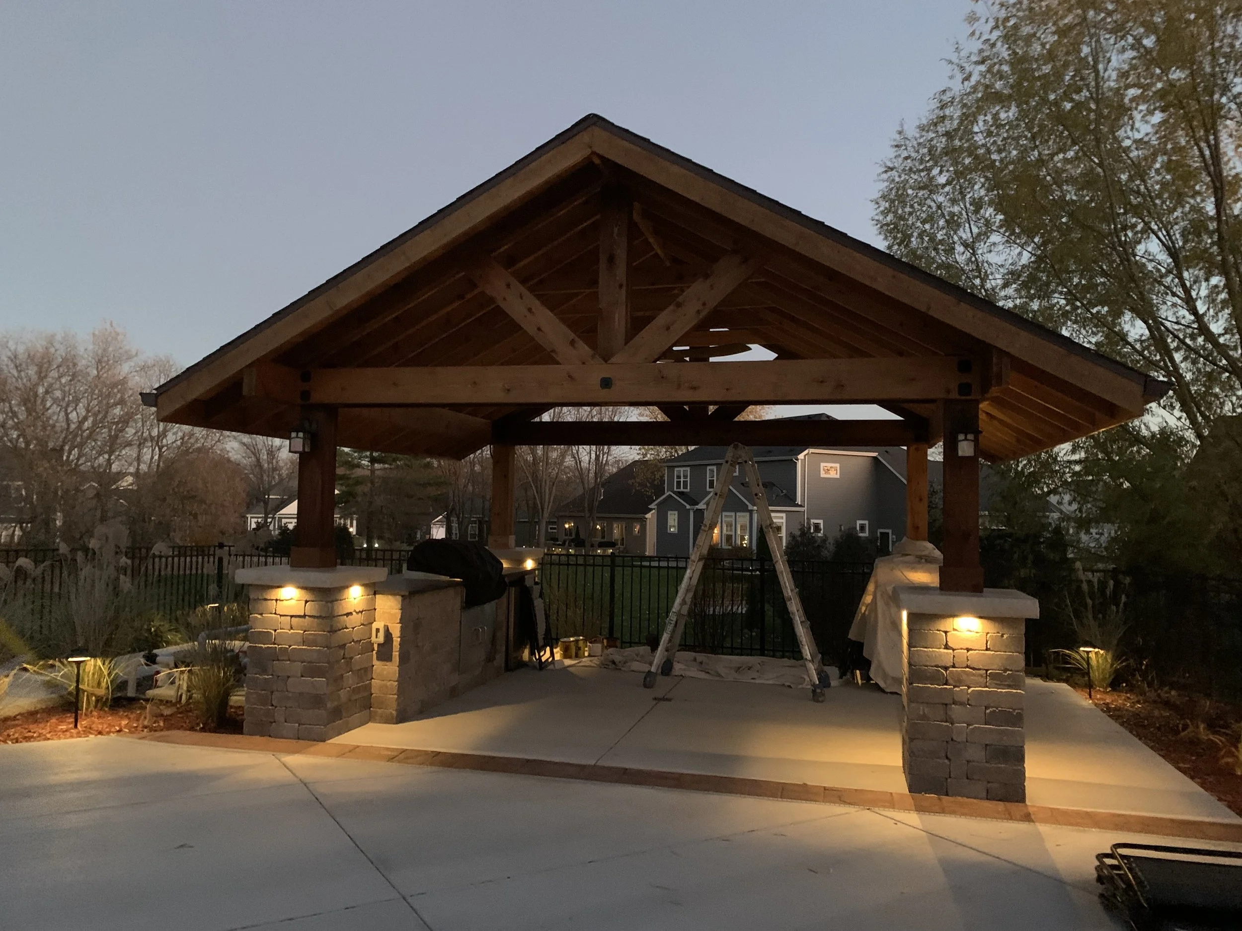 Outdoor pavilion with a gabled roof, brick pillars, and built-in lighting, under construction during evening with a ladder and tools inside.