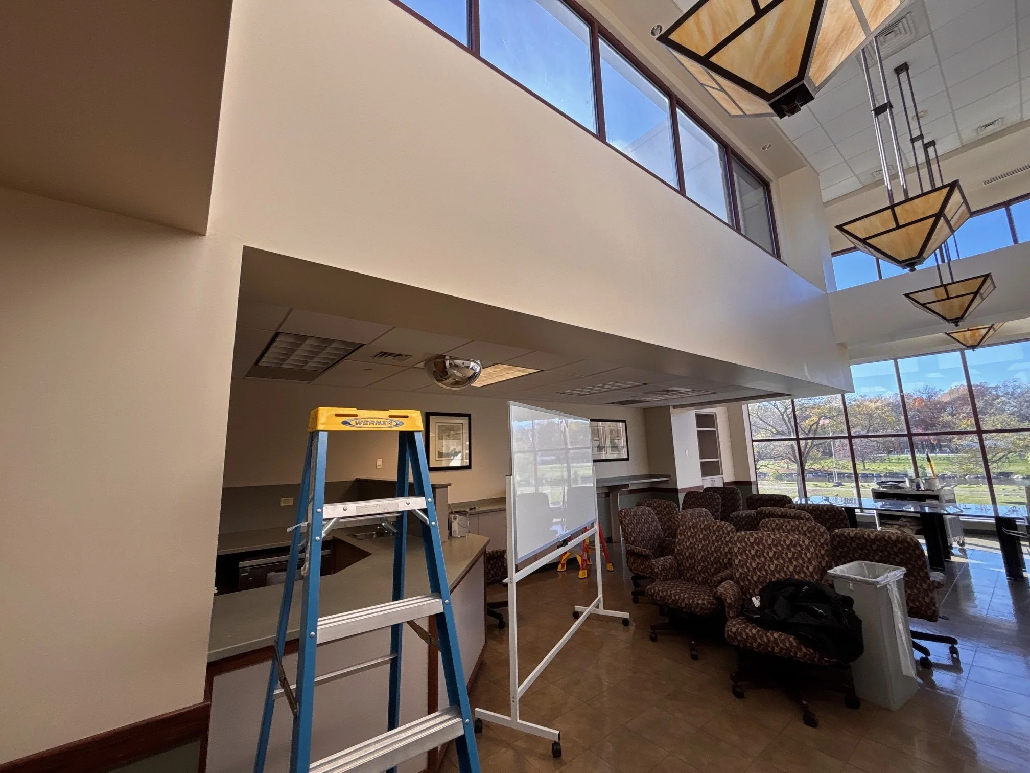 Interior of a room with large windows, a blue and yellow step ladder, chairs, a whiteboard, and ceiling lights.