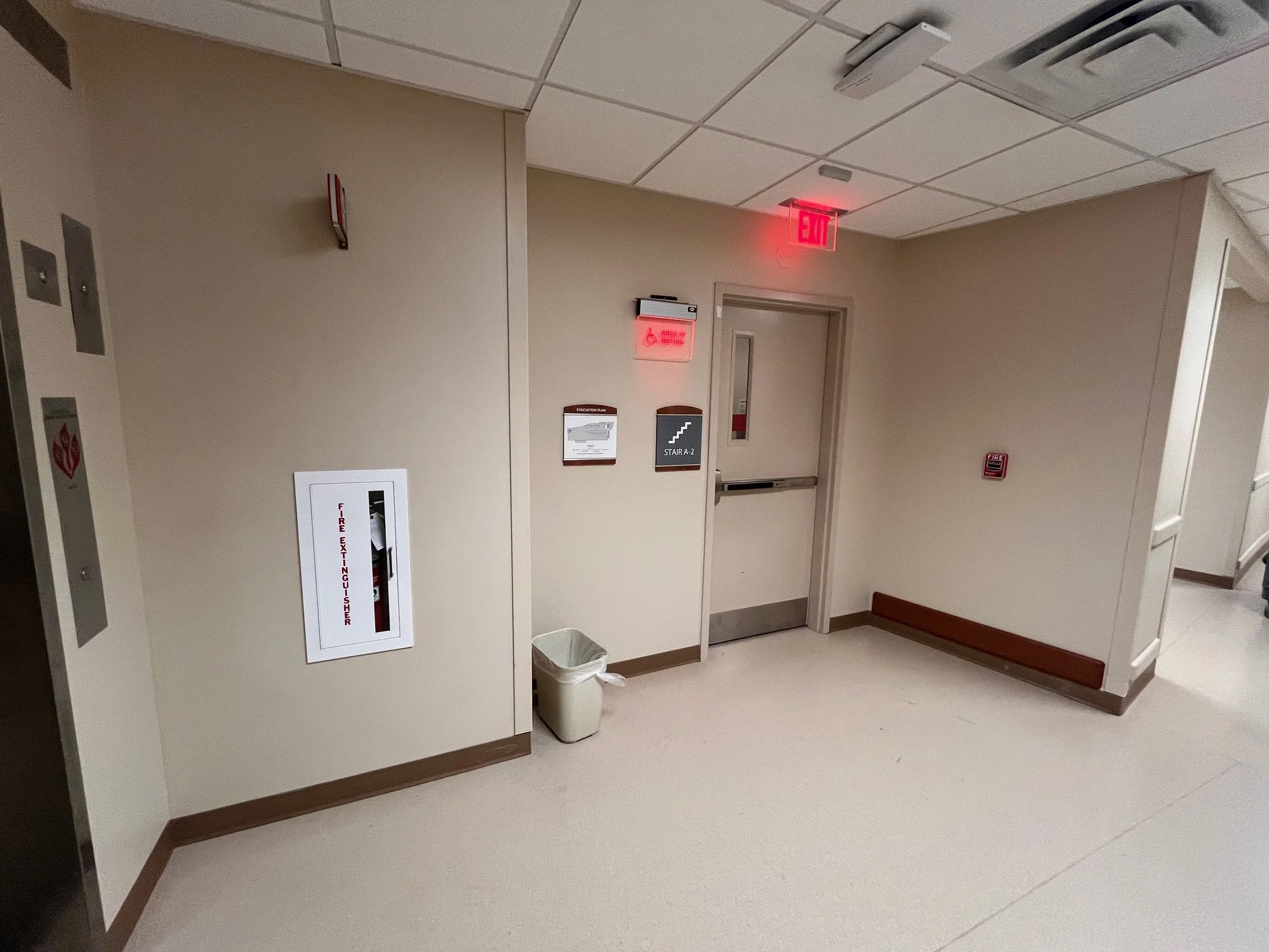 Hospital corridor with a fire extinguisher cabinet, a trash bin, an exit sign, and an emergency alarm. There is an elevator door and a stencil sign that says stair A2.