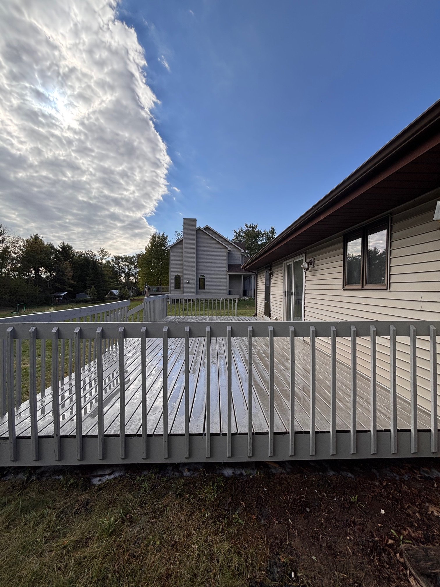 A wooden deck attached to a house, with a railing, in a residential area with trees and another house in the background. The sky is partly cloudy with some blue sky visible.