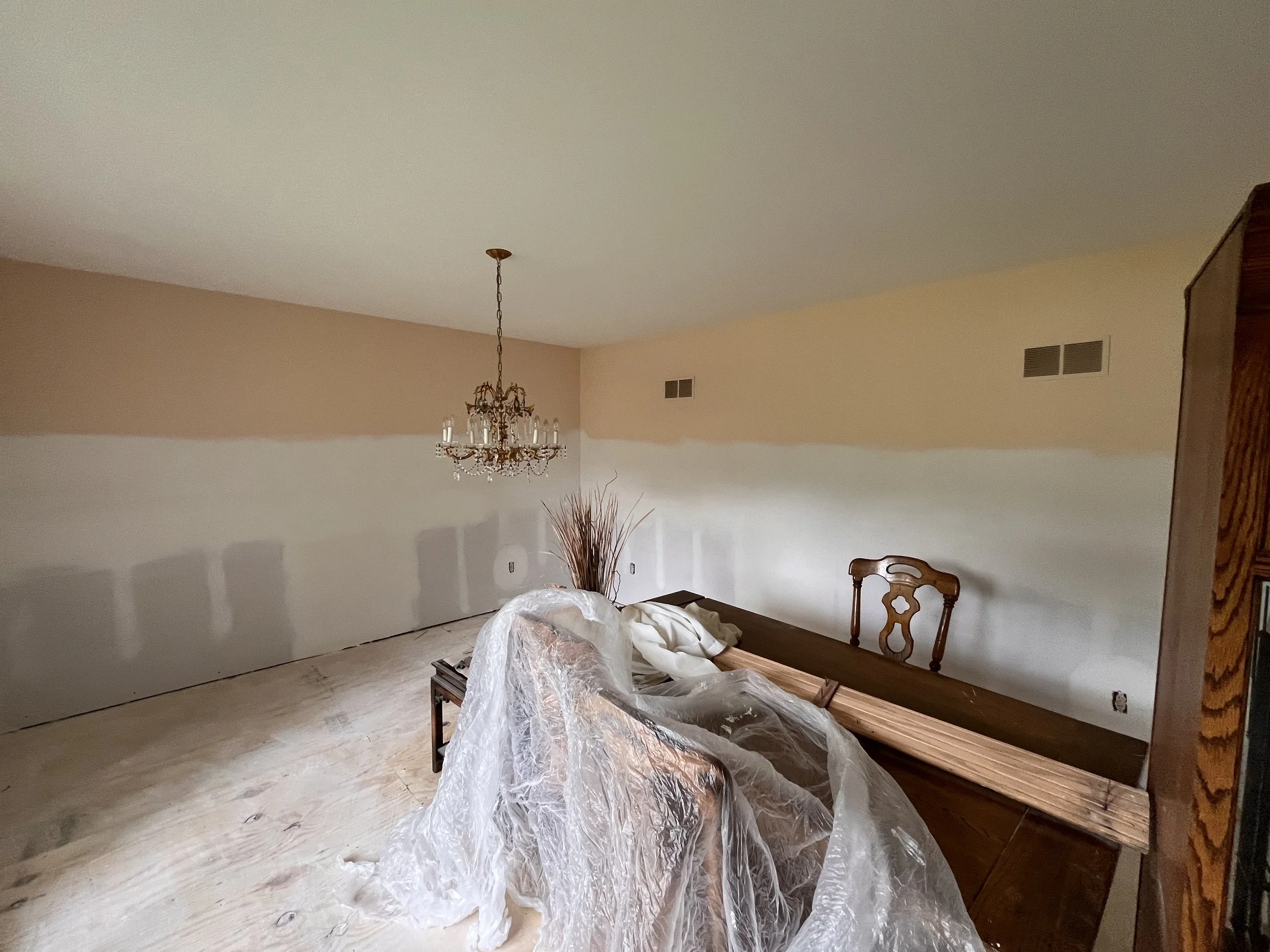 Living room undergoing renovation with a partially painted beige wall, a chandelier hanging from the ceiling, a wooden table, a chair, and plastic sheeting covering furniture.