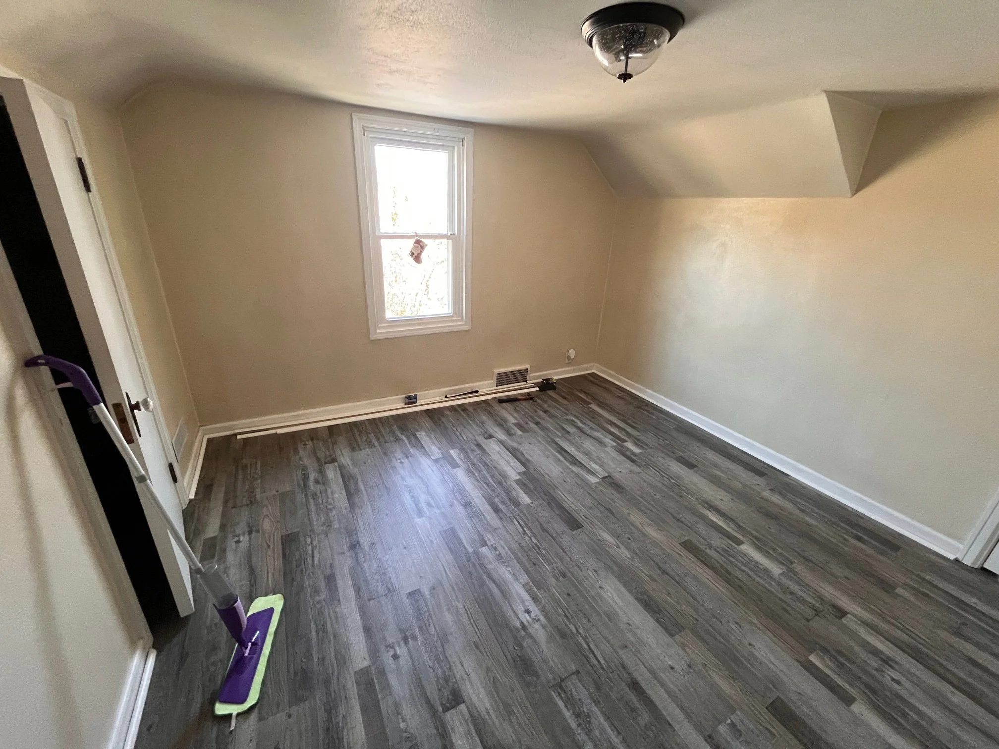 Empty room with wood flooring, beige walls, a window with a small hanging object, and a ceiling light fixture. There is a mop leaning against the wall.